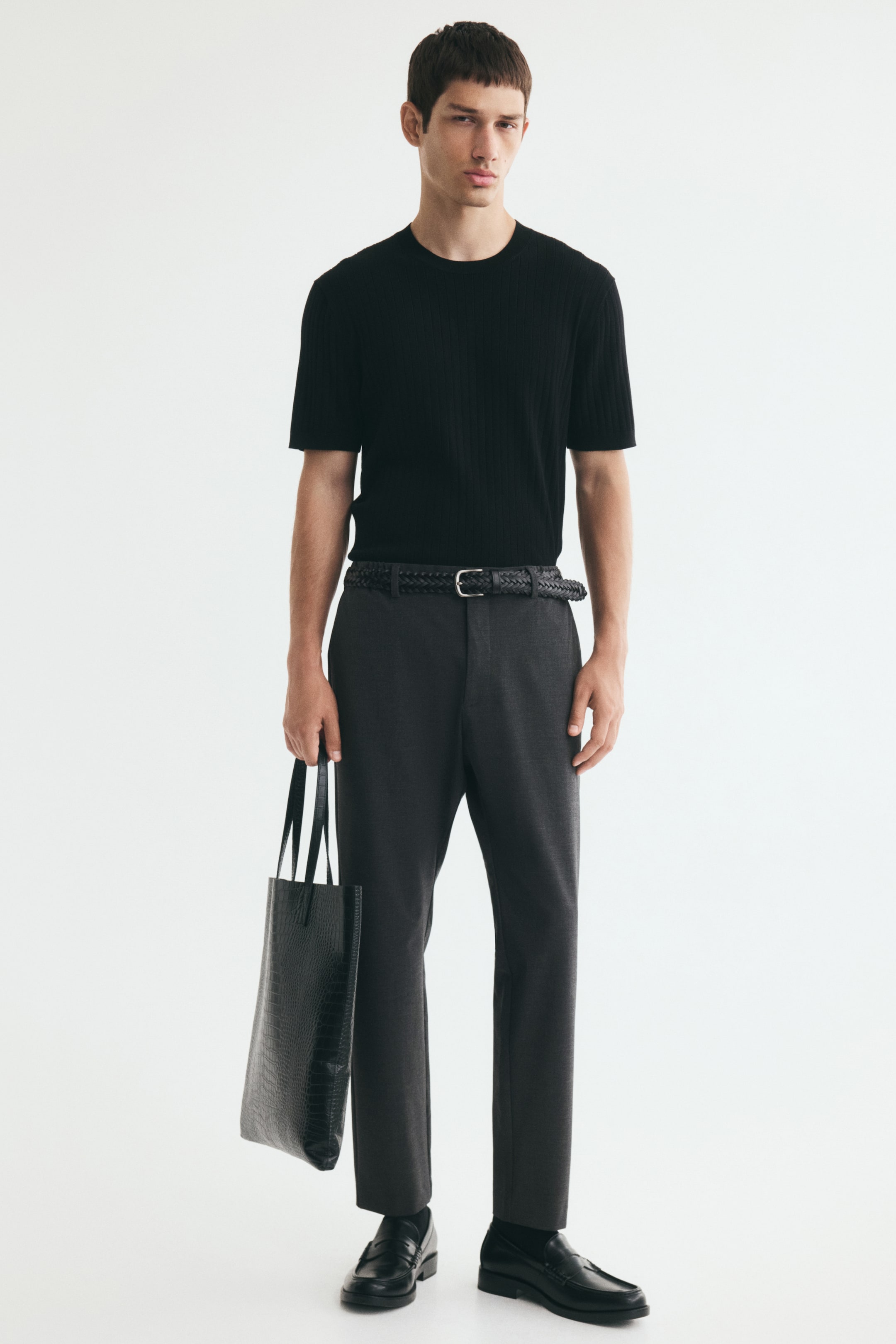 Büyük resmi görüntüle: Modelin üzerinde koyu gri renkli, ayak bileği boyunda chino pantolonlar yer alıyor; çapraz yan cepleriyle slim fit silueti öne çıkıyor. Pantolonlar, örgü siyah bir kemer, içine sokulmuş kısa kollu siyah bir tişört ve siyah loafer’larla kombinlenmiş; model ayrıca siyah dokulu bir tote çanta tutuyor.