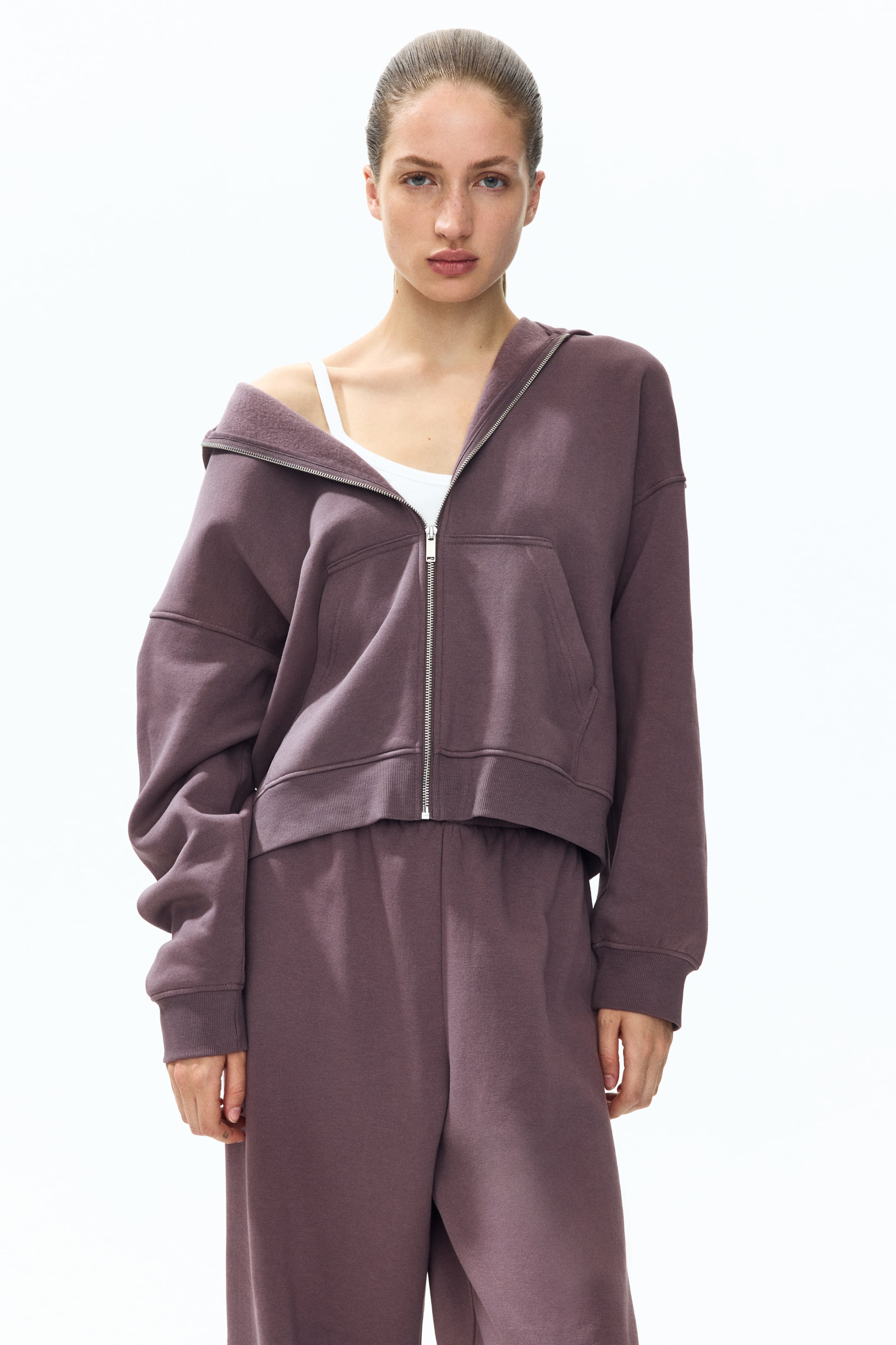 View larger image: A dark mauve loose-fit hoodie featuring a lined hood, zippered front, open front pockets, dropped shoulders, long sleeves, and ribbed cuffs and hem is showcased by the model. The model is styled with the hoodie partially unzipped, one shoulder casually draped over a white tank top, complemented by matching dark mauve sweatpants.