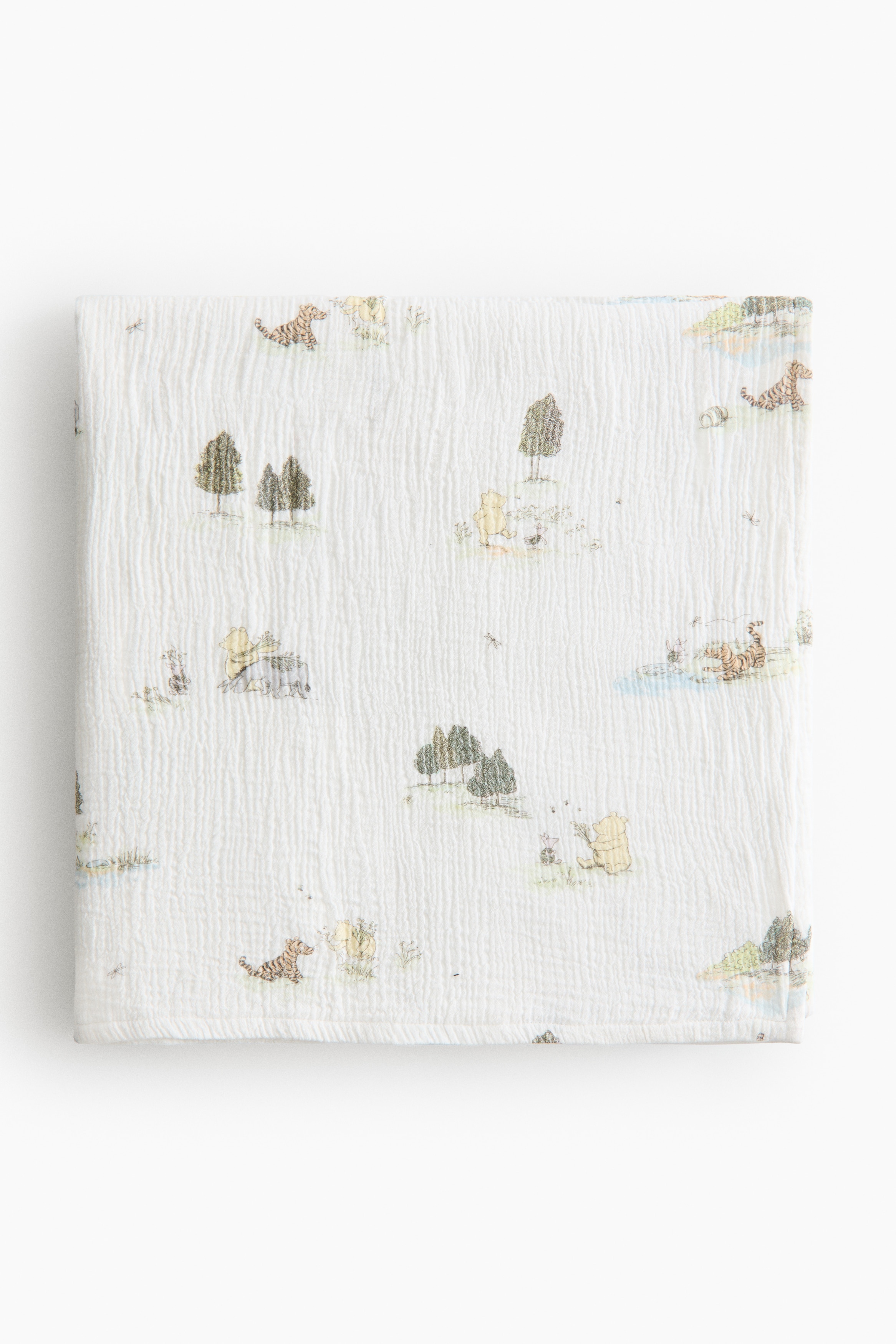 A greenish-yellow pear is resting upright, displaying its speckled skin and a brown stem extending from its top. It is positioned on a white, crinkled textile featuring subtle patterns of trees and animals, which is layered over a light green gingham textile.