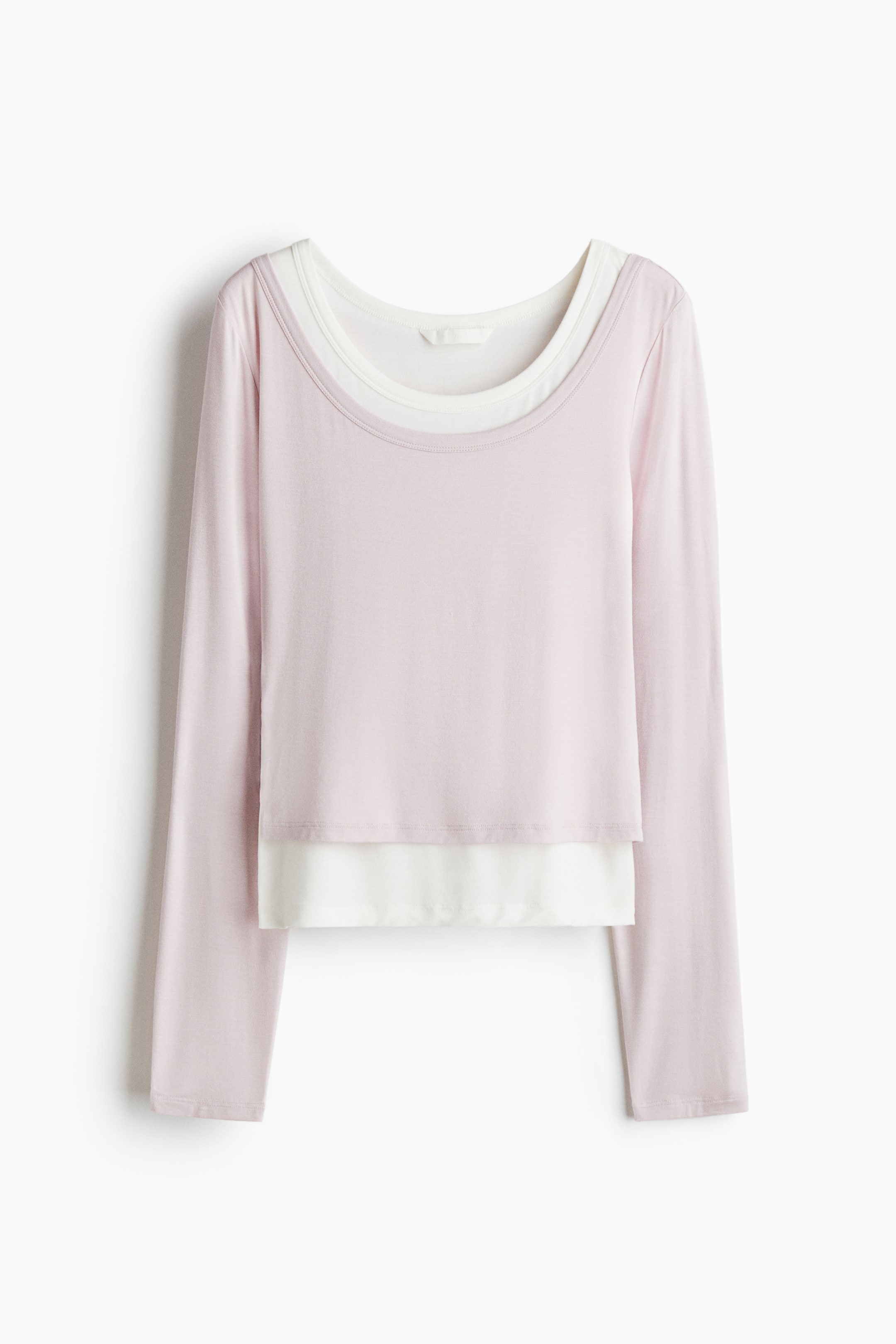 拡大画像を表示: 淡いピンクの長袖トップスは、前面にレイヤードデザインを見せています。丸いネックラインで白いアンダーレイヤーが見え、ピンクのトップスの短い裾の下に伸びています。
