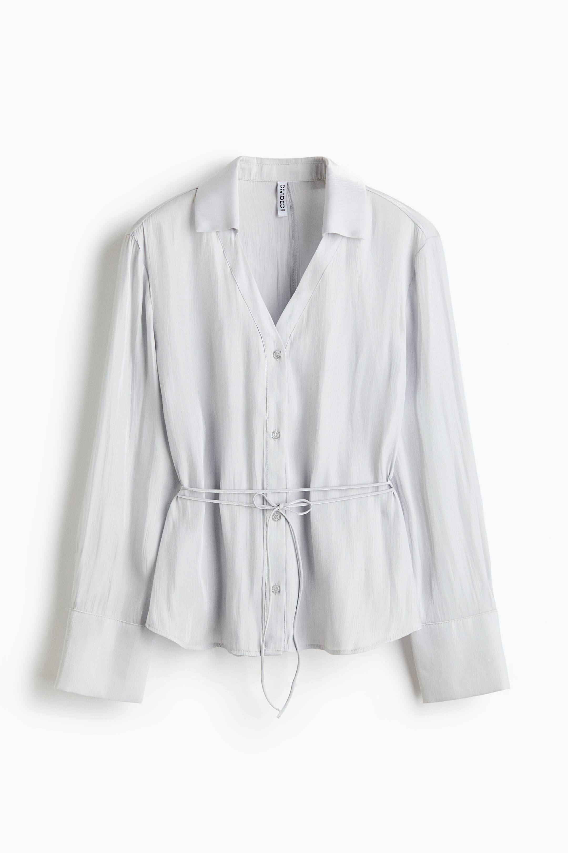 View larger image: A light grey long-sleeved blouse is presented facing the front, featuring a deep V-neckline with a resort collar and a button-down front. The blouse showcases wide cuffs on the long sleeves and a thin, matching fabric drawstring tie cinched at the waist.