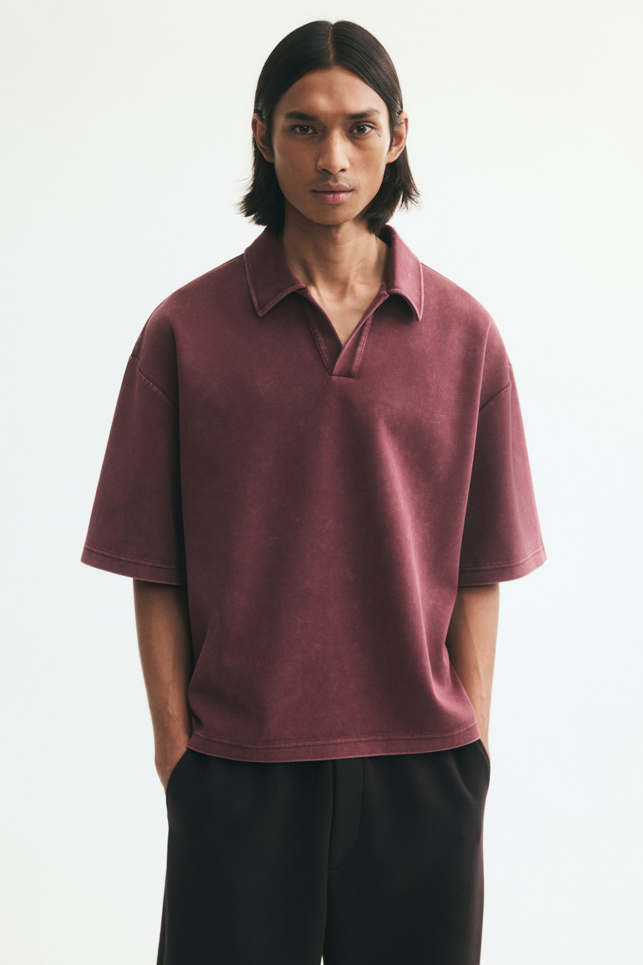 View larger image: A short-sleeve polo shirt in washed burgundy is worn by the model. The shirt features an oversized silhouette with dropped shoulders, an open collar, and a straight hem, paired with dark trousers.