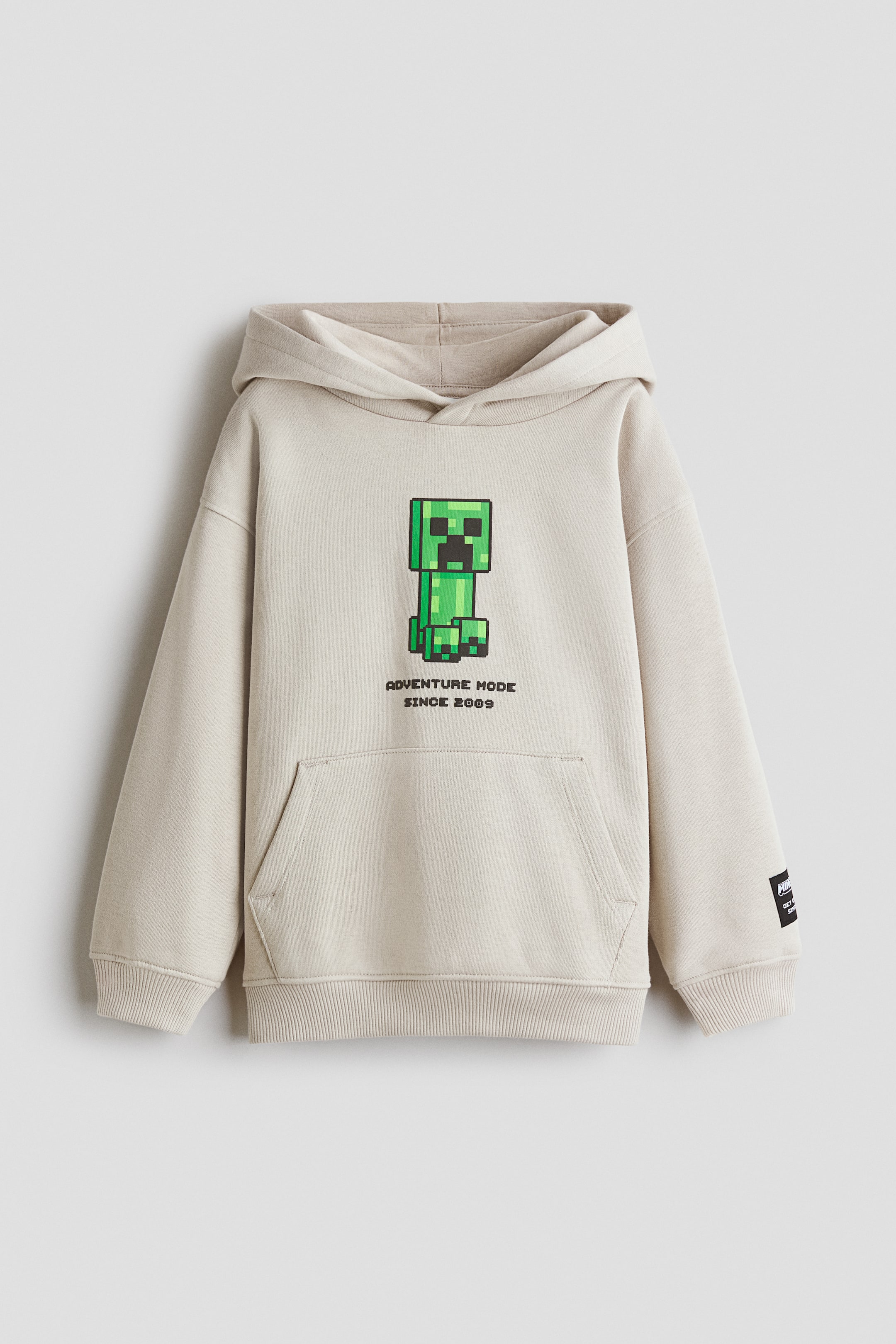 Vezi imaginea mai mare: Produsul este un hanorac cu croială lejeră, cu față frontală, din material fleece tip sweatshirt, de culoare bej deschis. Are imprimat pe piept un personaj Creeper pixelat verde și textul „ADVENTURE MODE SINCE 2009”, precum și o glugă căptușită cu jerseu și panou frontal suprapus, umeri căzuți, buzunar tip kangaroo și manșete și tiv striate.