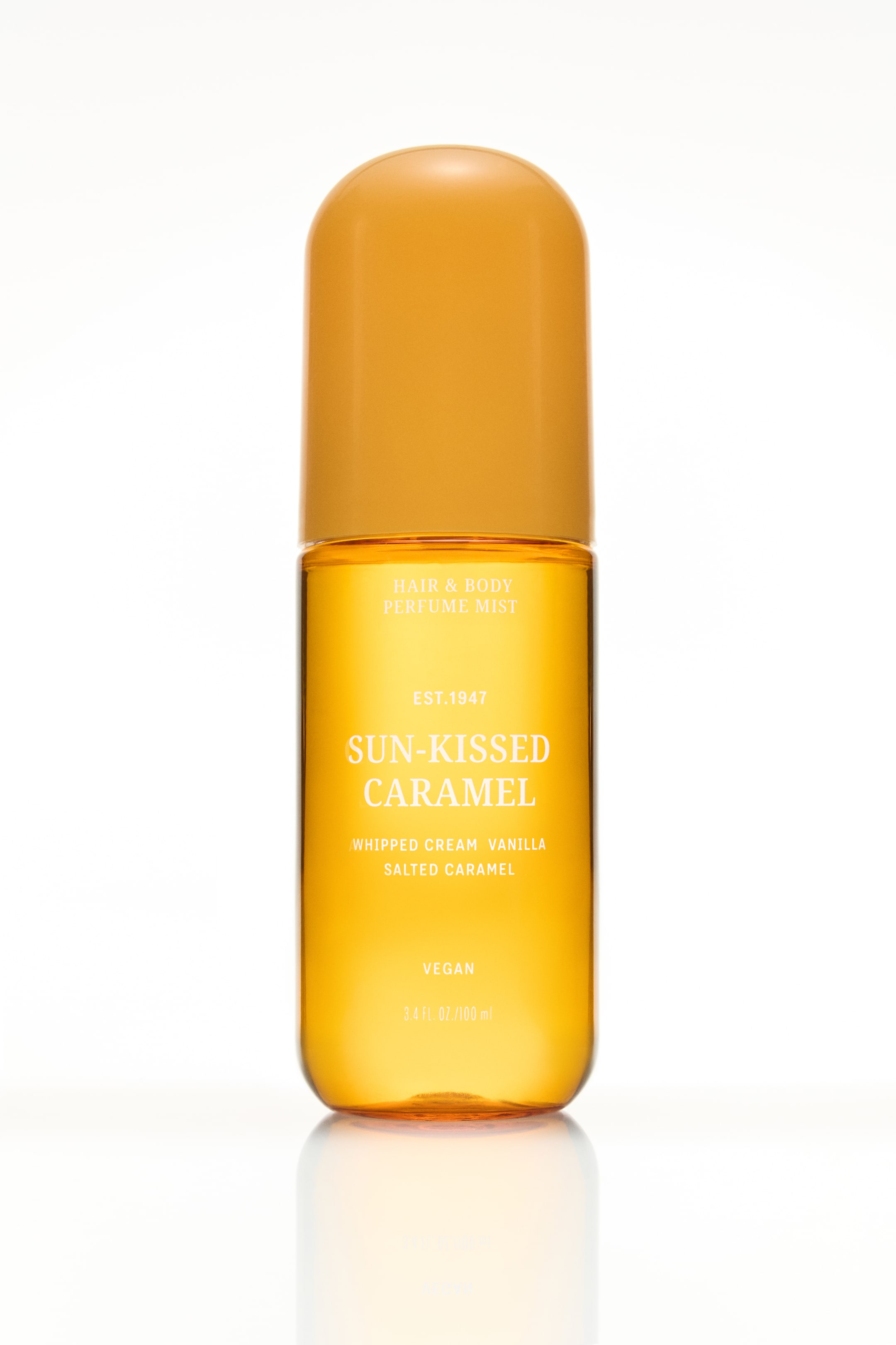 Ampliar la imagen: Una botella de bruma perfumada para cabello y cuerpo está de pie, mirando hacia adelante. Tiene un cuerpo cilíndrico transparente lleno de líquido color ámbar y una tapa redondeada opaca del mismo tono ámbar. El frente de la botella muestra texto blanco, incluyendo 'Bruma Perfumada para Cabello y Cuerpo' y 'Caramelo Besado por el Sol'.
