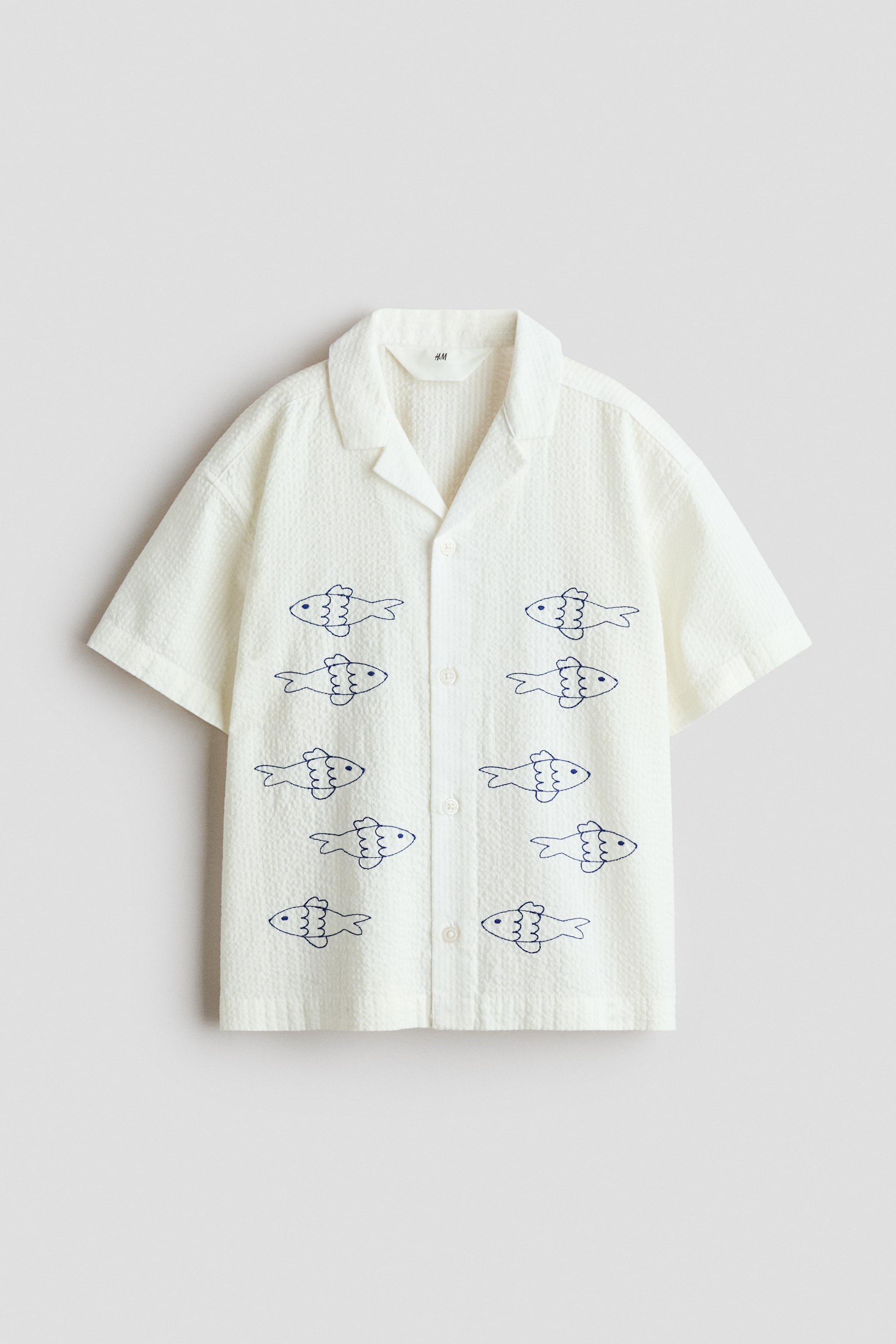 View larger image: A relaxed-fit shirt crafted from soft cotton seersucker, showcasing embroidered blue fish accents on the front. It features a resort collar, button-down front, dropped shoulders, and short sleeves.