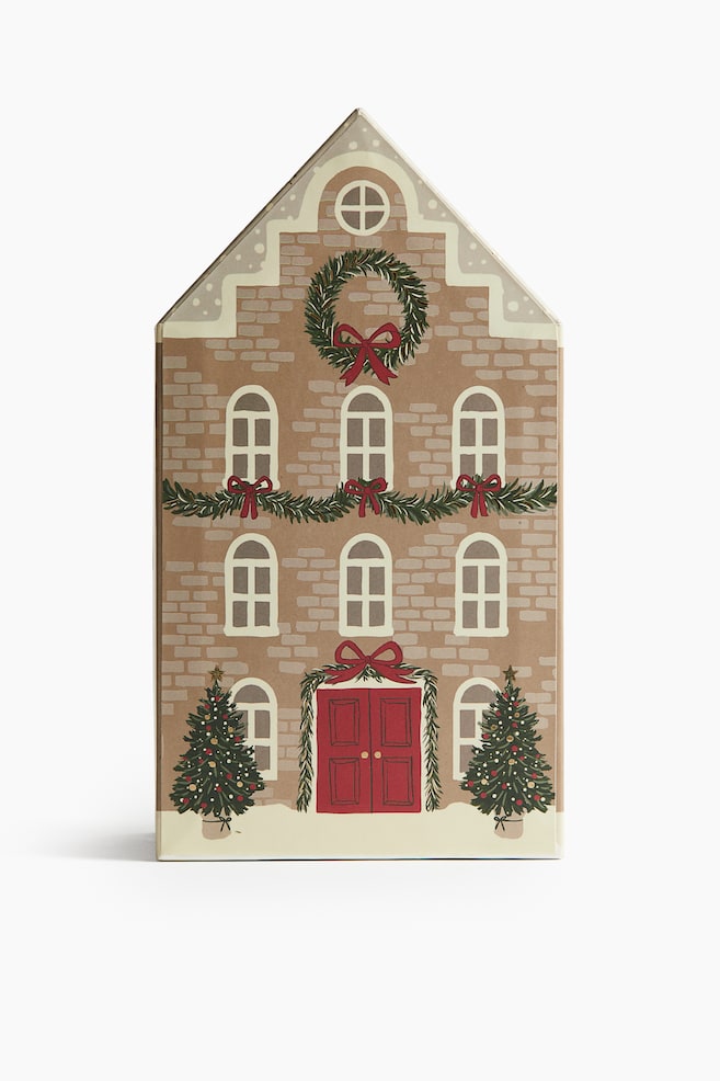En framsida av en husformad presentlåda har en brun tegelstensdesign med vita välvda fönster, en röd dubbeldörr och två gröna julgranar. Lådan är också dekorerad med gröna girlander och kransar prydda med röda rosetter. - 1