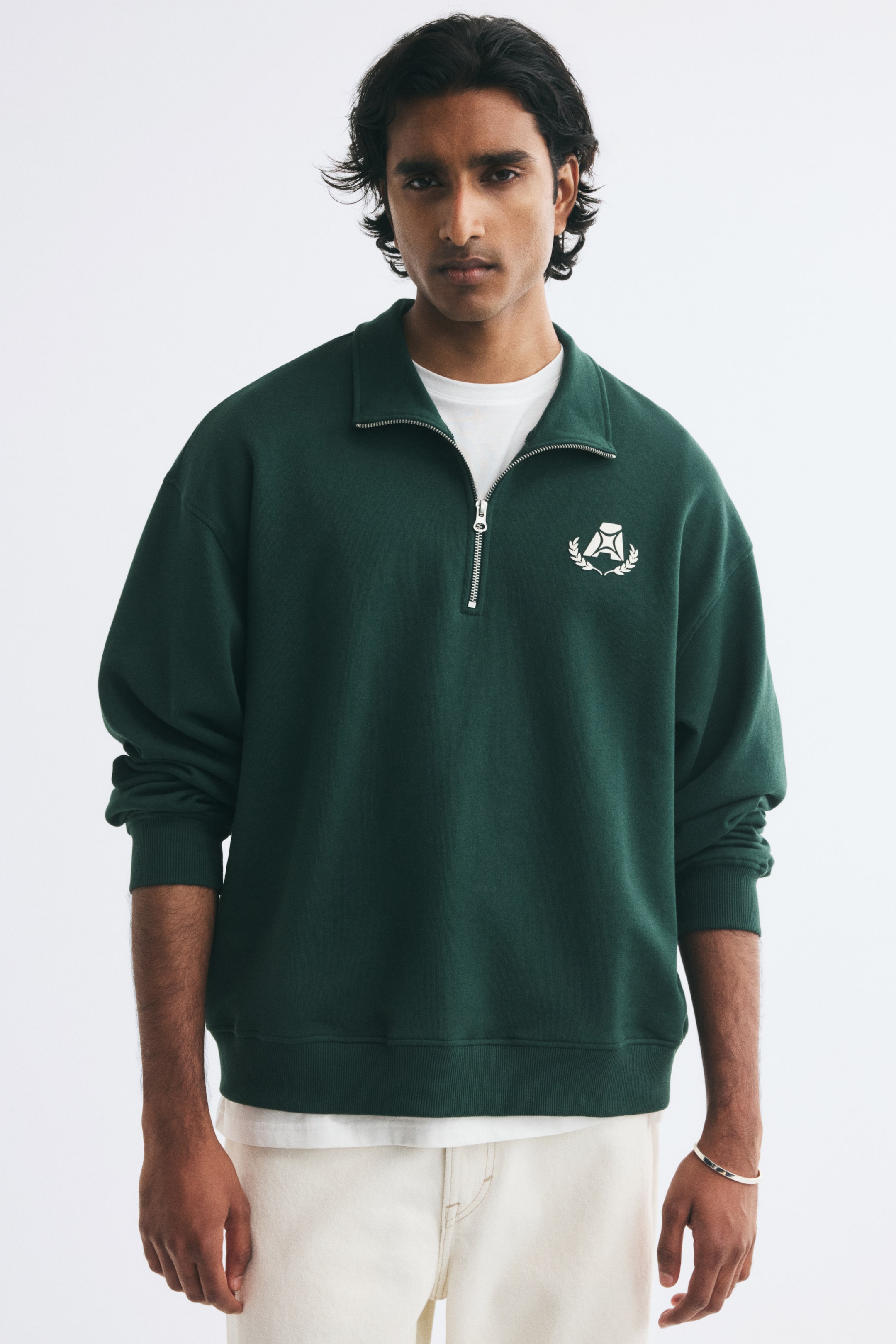View larger image: The model is wearing a dark green half-zip sweatshirt over a layered white T-shirt, paired with off-white trousers. The sweatshirt features a partially unzipped collar, ribbed cuffs and hem, and a small white graphic logo on the left chest.