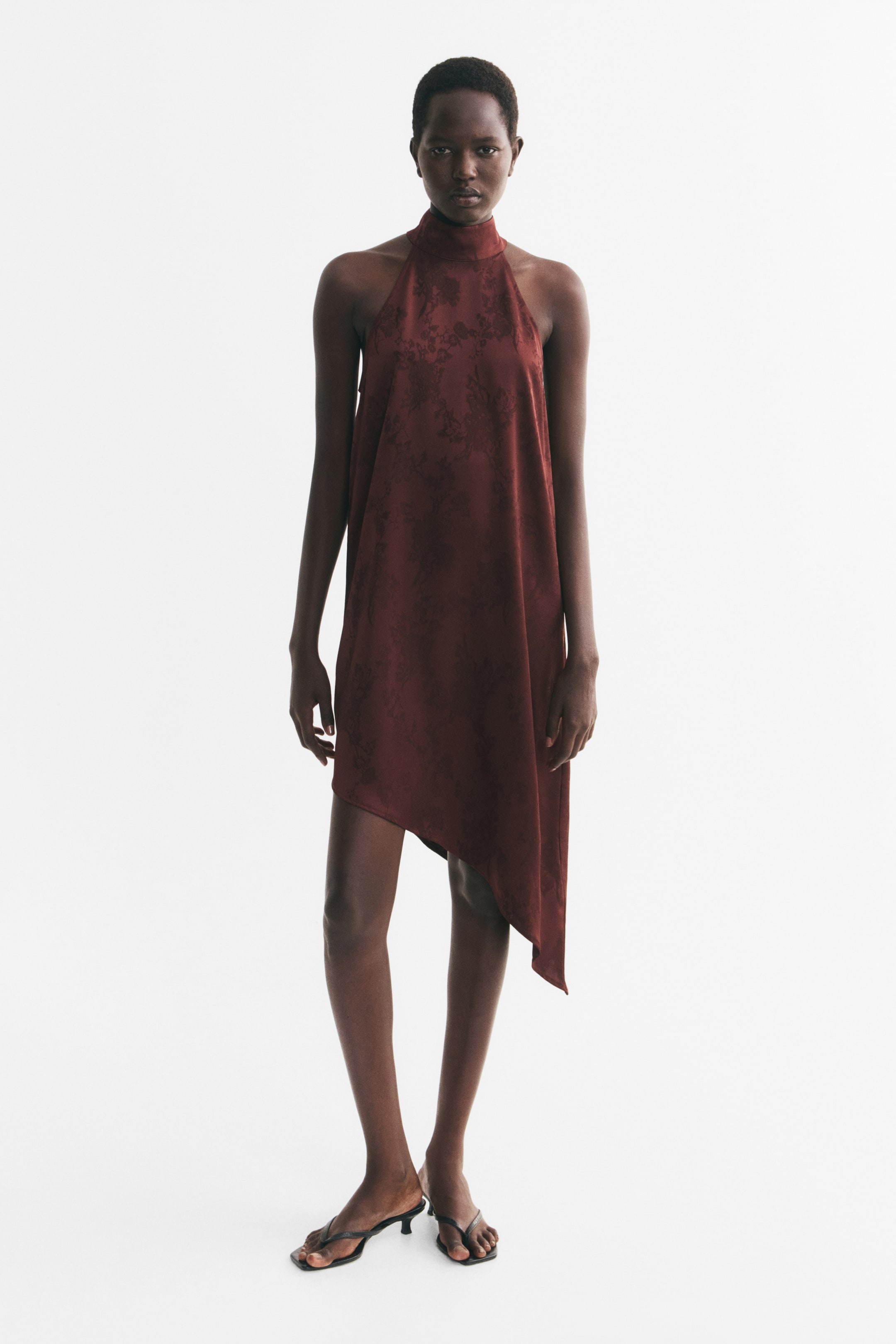Ampliar la imagen: La modelo lleva un vestido rojo oscuro de cuello halter con un sutil patrón tonal y un dobladillo asimétrico, además de sandalias negras tipo thong con tacón bajo tipo kitten heel.
