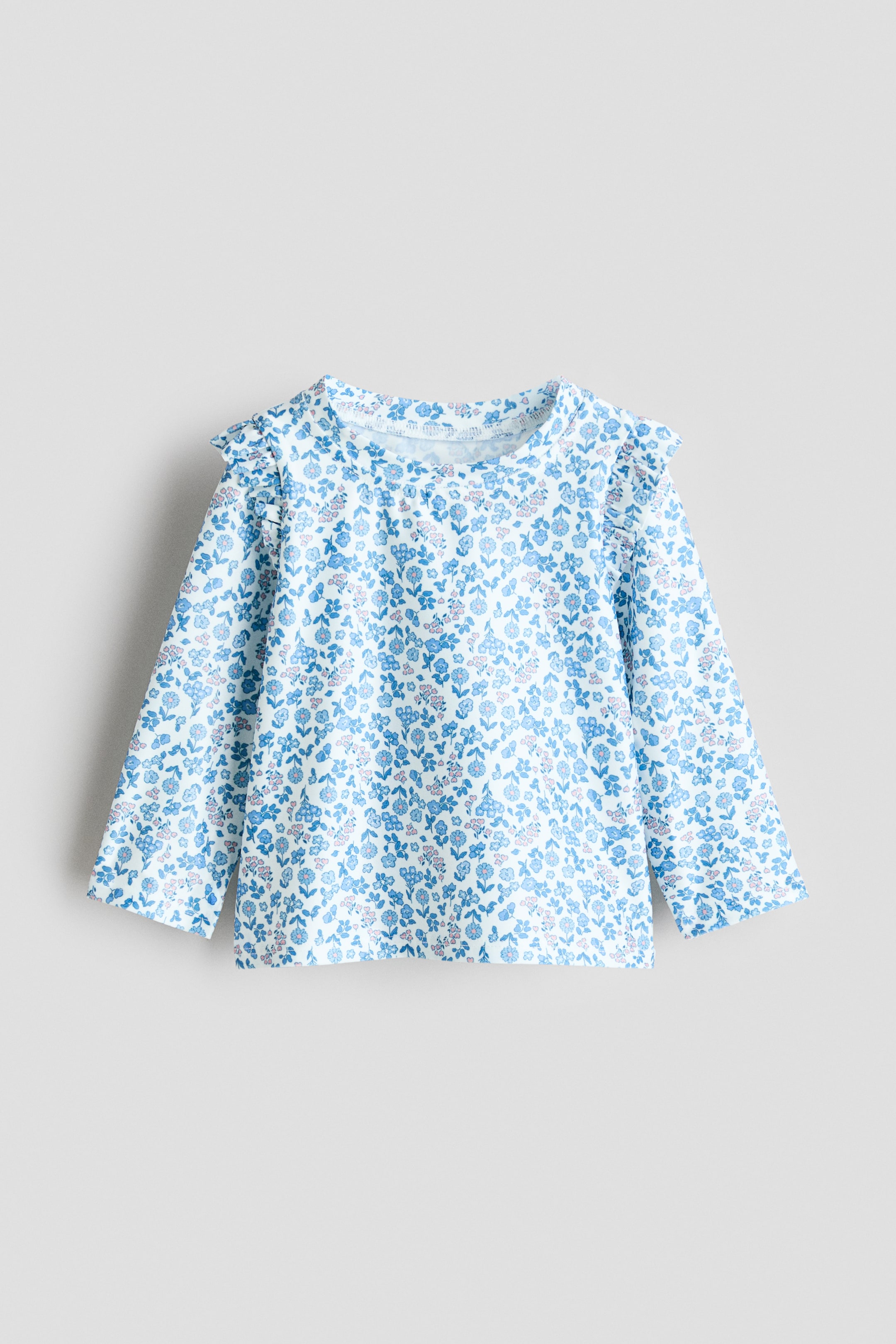 Agrandir l'image: Un haut de bain avec protection UV est montré de FACE, présentant un motif intégral de fleurs bleues et roses claires avec des feuilles vertes sur un fond blanc, conçu avec un col rond, des manches longues et des détails à volants sur les épaules.