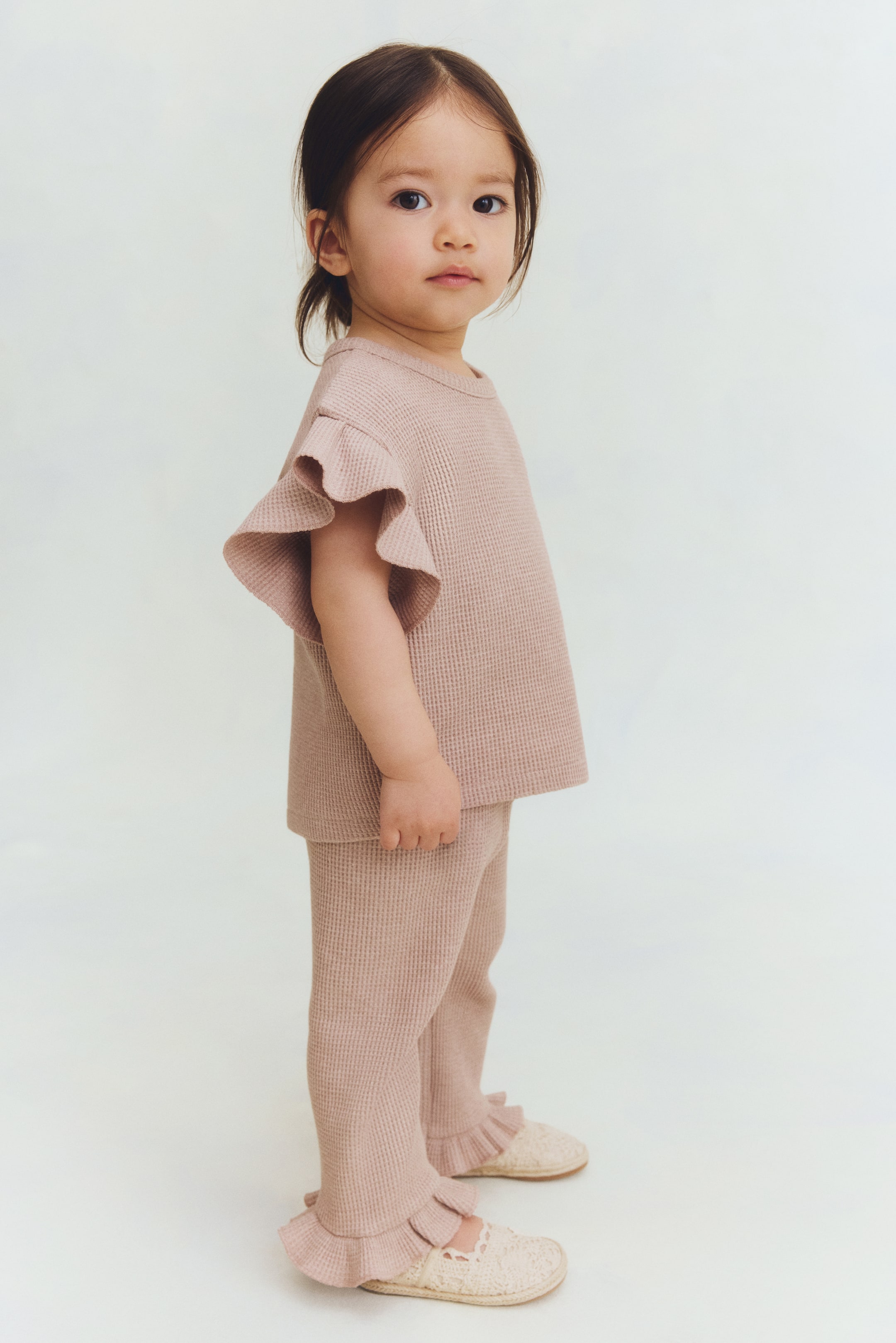 View larger image: A light pink two-piece waffle knit outfit is showcased from a three-quarter front angle, featuring a top with a crew neck and ruffled short sleeves, paired with matching long pants that have ruffled ankle hems.