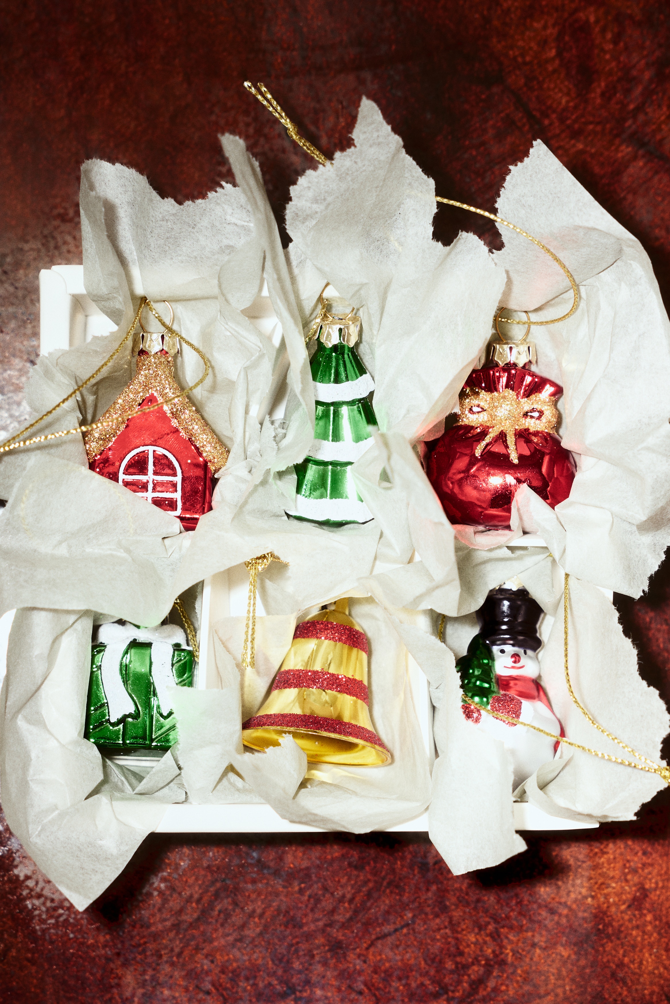 Les décorations de Noël en plastique présentent divers motifs, chacun doté d'un œillet en haut avec un cordon doré scintillant. Le DEVANT des décorations montre un sac cadeau rouge brillant avec un nœud doré, un bonhomme de neige tenant un petit arbre vert, un sapin de Noël vert à étages avec des détails blancs, une maison rouge brillante avec un toit marron scintillant et un cadre de fenêtre blanc, une cloche dorée avec des rayures horizontales rouges, et une boîte cadeau verte brillante avec un ruban et un nœud blancs.