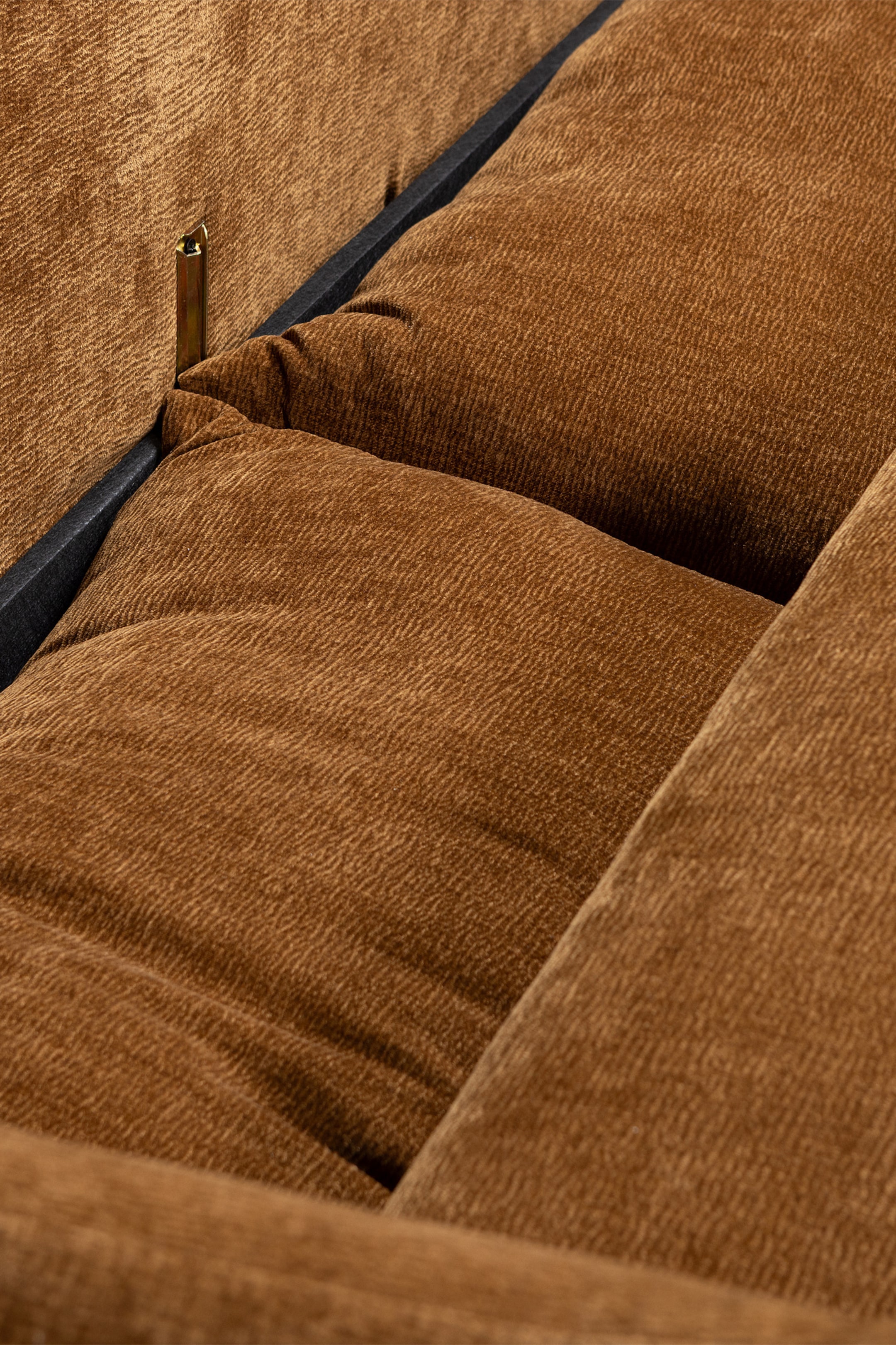 Grotere afbeelding bekijken: Dik gevoerde kussens in een warmbruine, horizontale geribbelde stof vormen de zitting en rugleuning van een meubelstuk. Een donker, mogelijk zwart, frame scheidt de kussens, en een kleine metalen beugel verbindt de rugleuning met het frame.