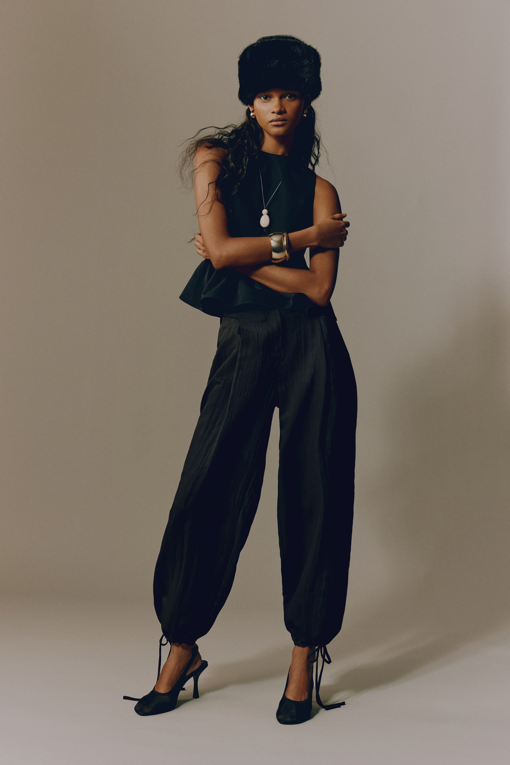 Größeres Bild anzeigen: Eine Person trägt ein schwarzes, ärmelloses Top mit hohem Kragen und Schößchen-Saum, kombiniert mit einer schwarzen, weit geschnittenen Hose mit Falten-Details und gerafften Bündchen mit Kordelzug. Dazu werden ein schwarzer Fellhut, eine lange Silberkette mit weißem, ovalem Anhänger, eine breite Armspange in Goldoptik und schwarze Slingback-Pumps getragen.