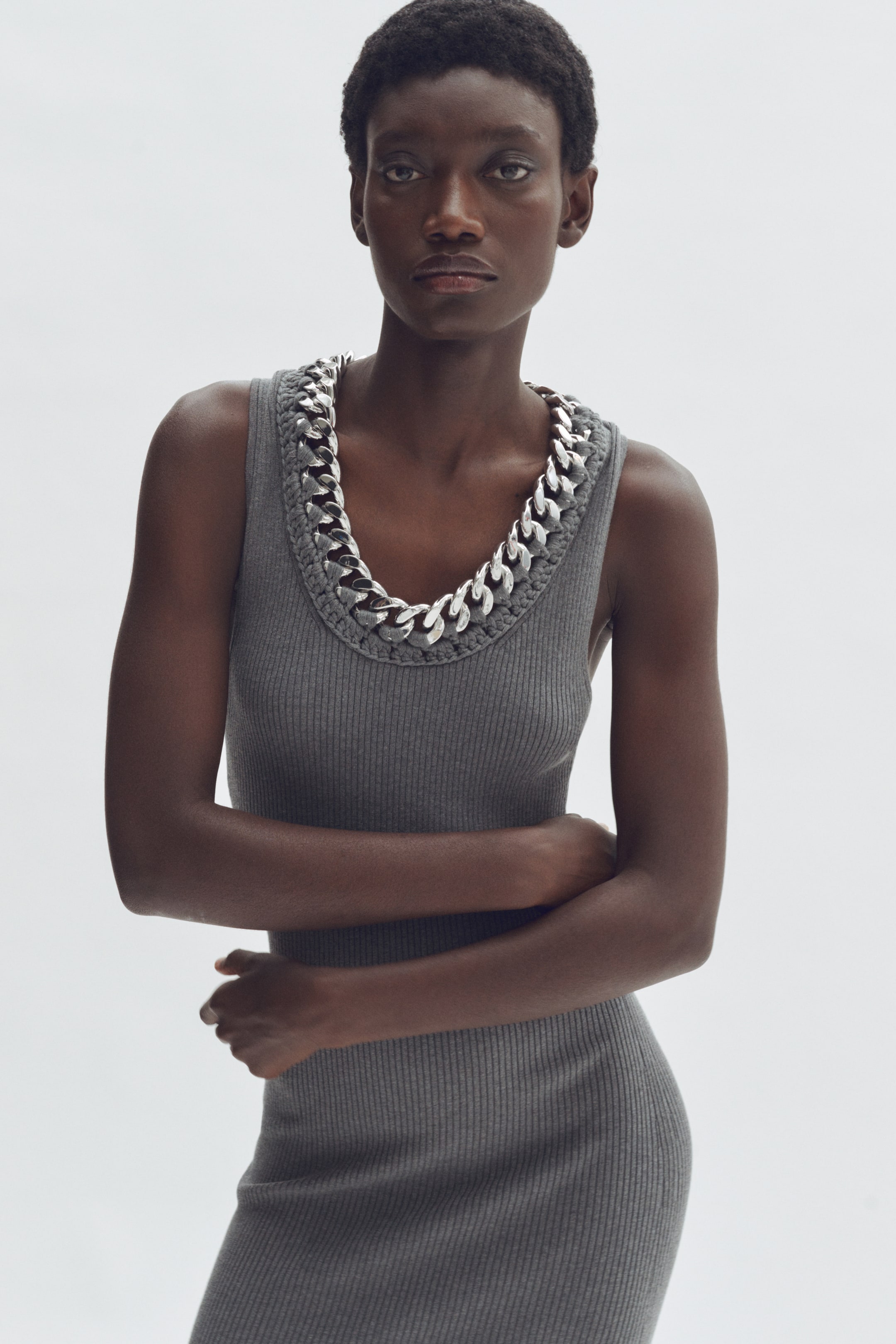 Nagyobb kép megtekintése: Egy szürke bordázott kötött ruha, amelyet egy nagy, ezüstszínű láncszemekkel átszőtt szegélyes kerek nyakkivágás jellemez, szorosan illeszkedik és elölről nézve van bemutatva.