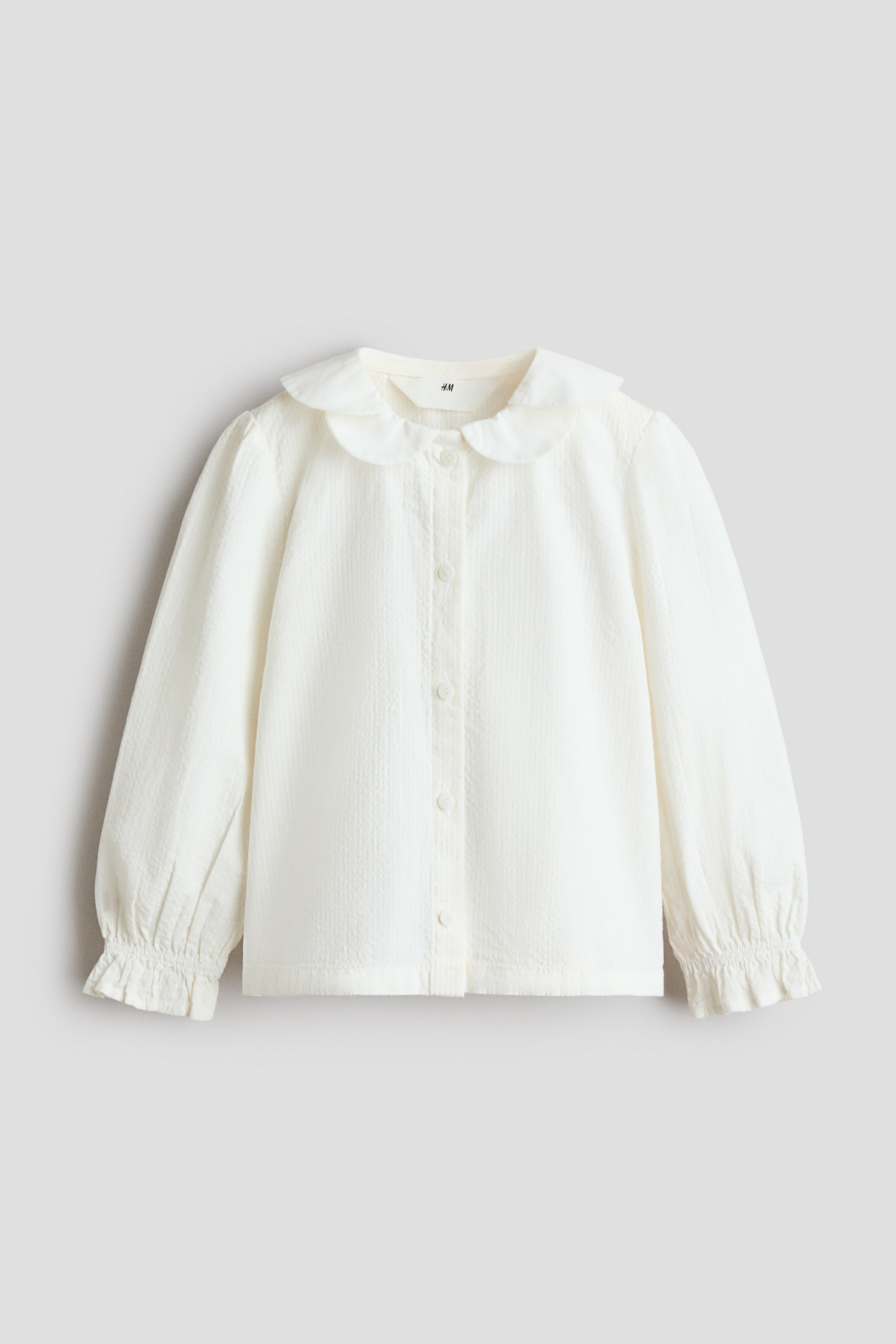 View larger image: A white blouse is shown front-facing, featuring a rounded Peter Pan collar and a subtle textured weave. It is designed with a button-down front closure and long sleeves ending in gathered, ruffled cuffs.