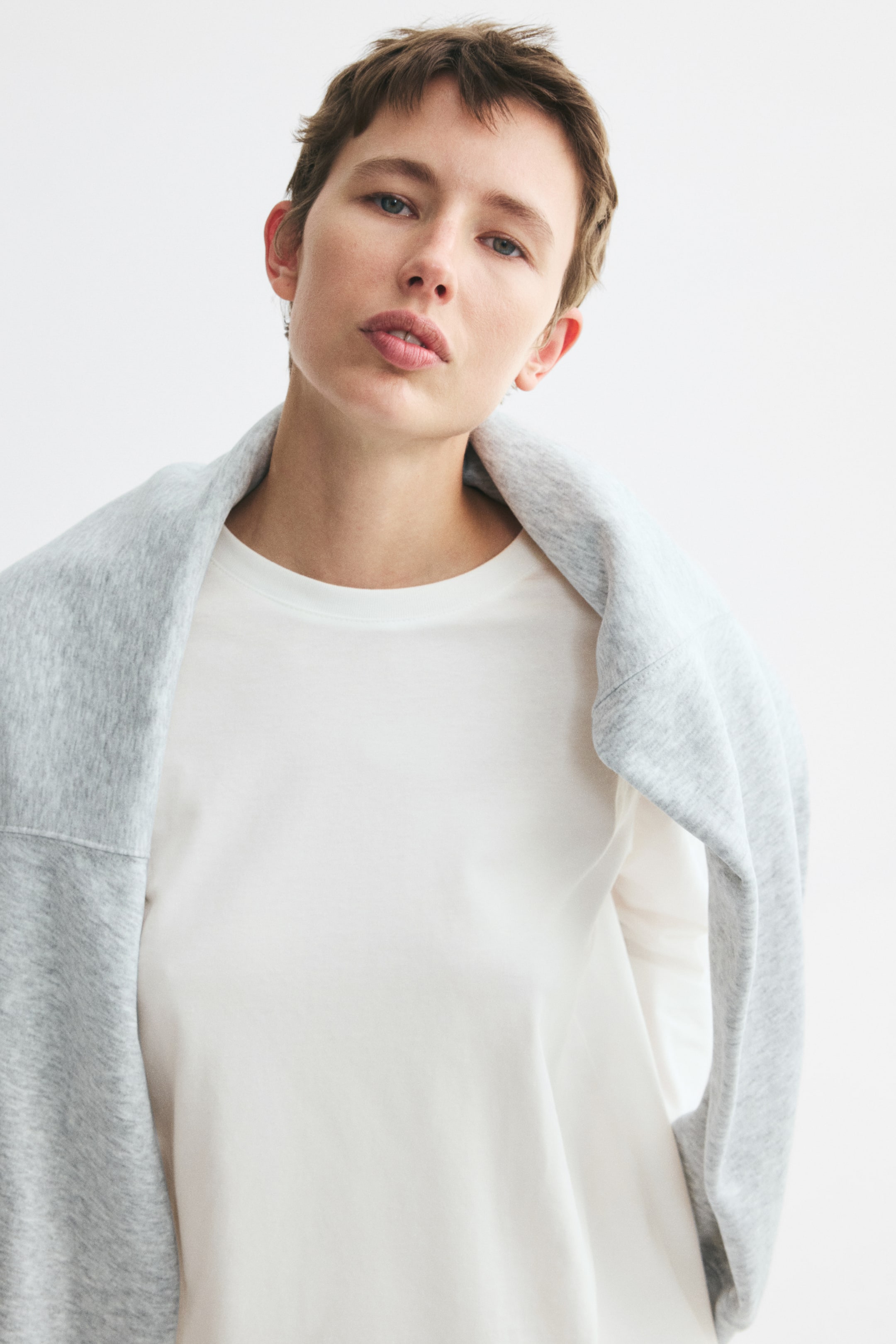 View larger image: The model is wearing a white long-sleeved cotton jersey top with a round neckline, with a grey sweatshirt draped over the model’s shoulders.