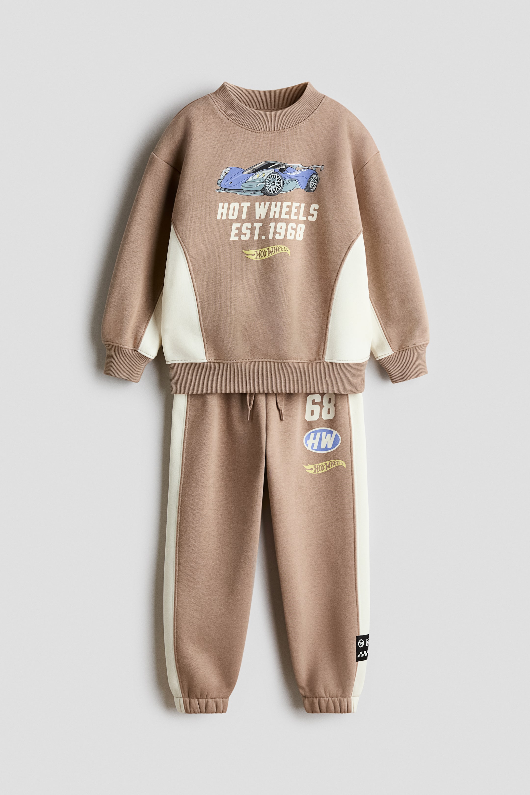 View larger image: A front-facing sweater and track pants set in light brown fleece is showcasing cream-coloured side panels and printed Hot Wheels graphics. The sweater features a round, rib-trimmed neckline with ribbed cuffs and hem, while the matching track pants feature an elasticated drawstring waist and elasticated hems.