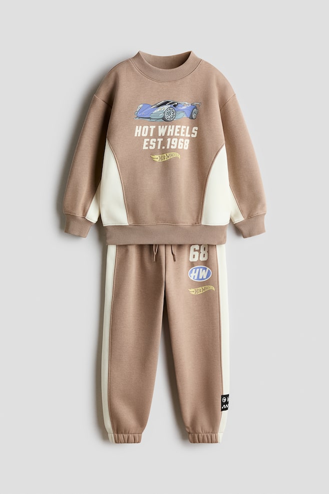 A front-facing sweater and track pants set in light brown fleece is displaying cream-colored side panels and printed Hot Wheels graphics. The sweater is showing a round, rib-trimmed neckline with ribbed cuffs and hem, while the matching track pants are displaying an elasticated drawstring waist and elasticated hems. - 1
