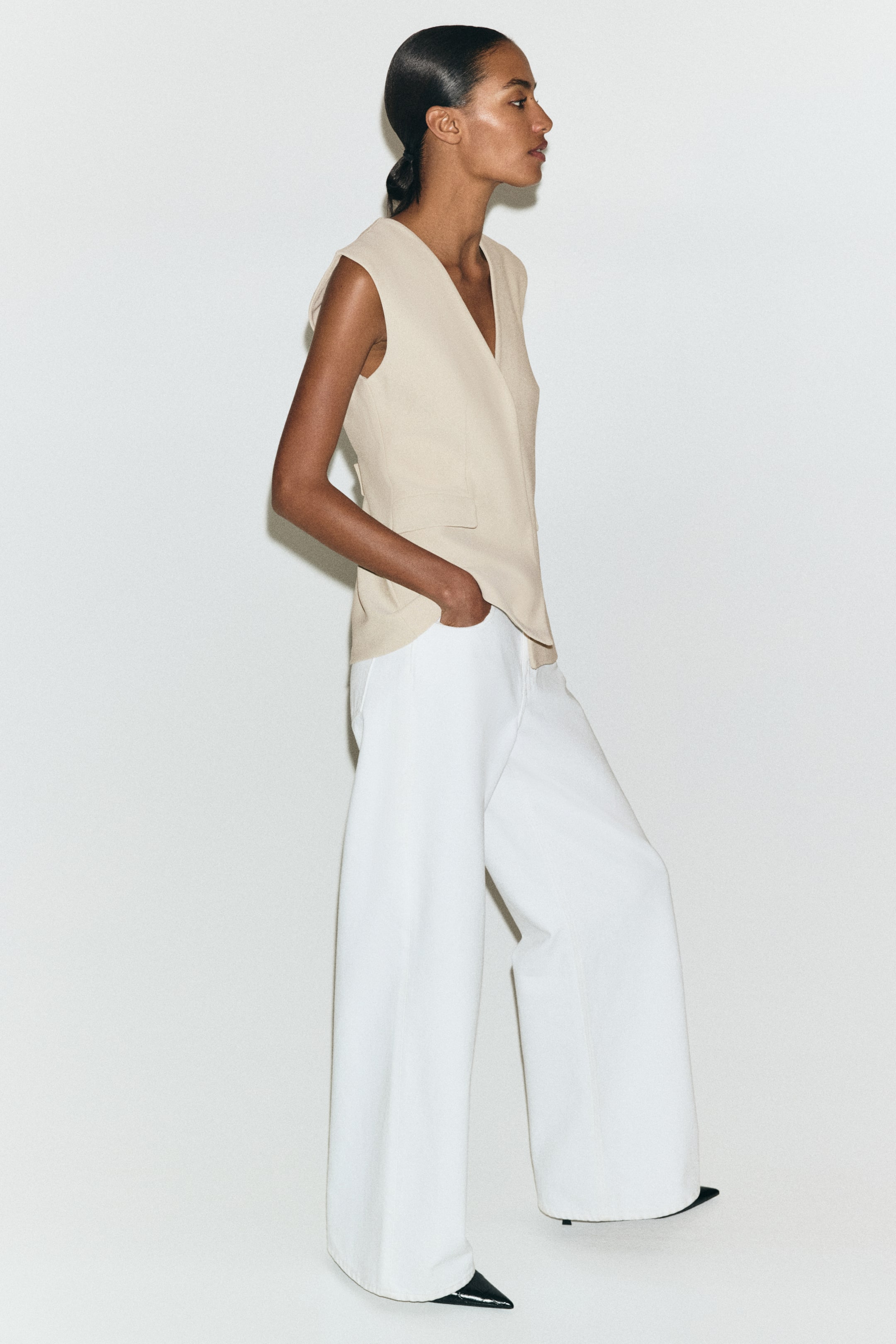 View larger image: A light beige sleeveless vest is presented from a side angle, featuring a V-neckline and an overlapping front design with a small flap pocket, being paired with white wide-leg pants that are flowing loosely.