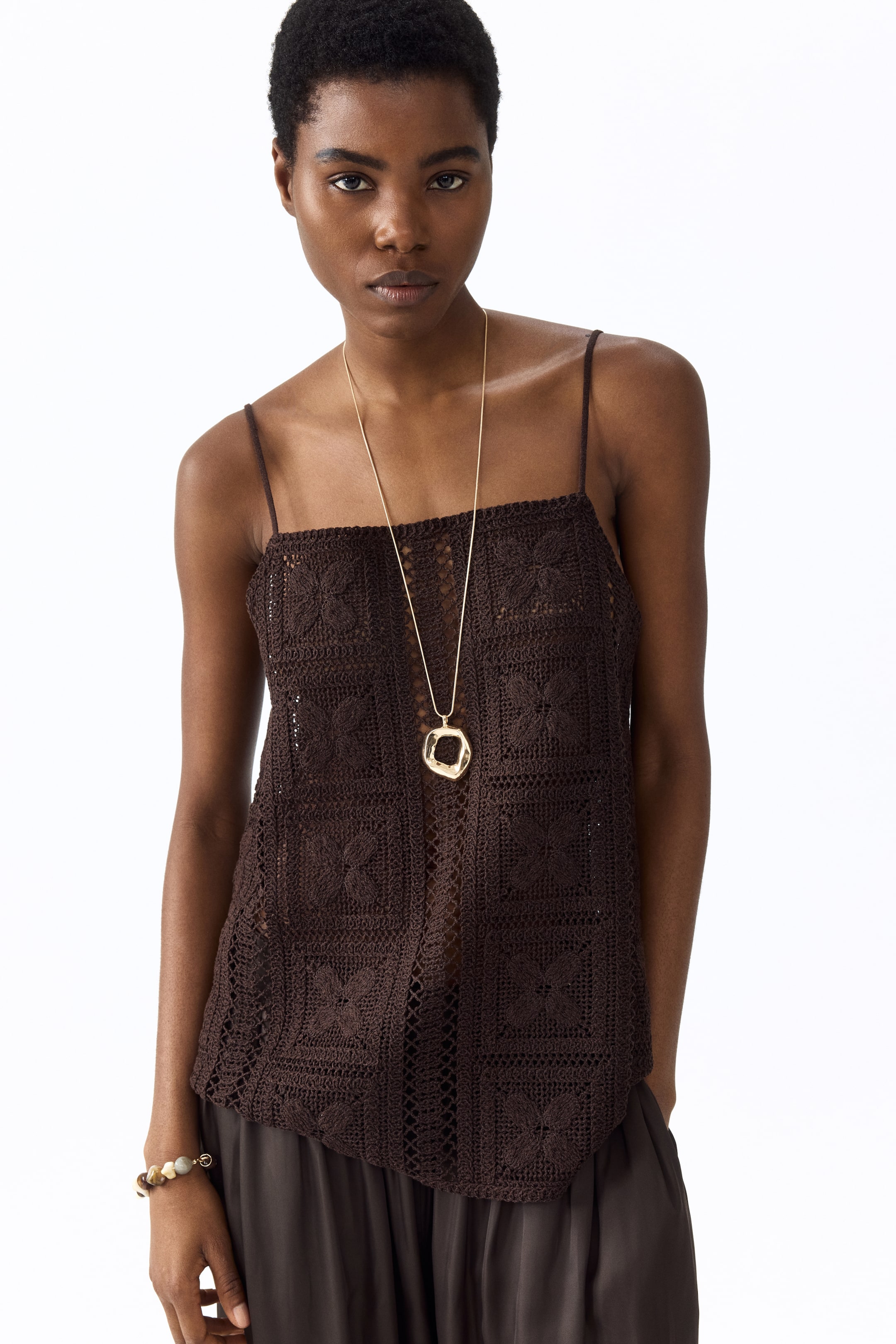 View larger image: A dark brown crochet top is being worn, featuring thin spaghetti straps and a square neckline. It displays an open-knit pattern of connected squares, each containing a floral design, and is styled with a long gold necklace and dark brown trousers.