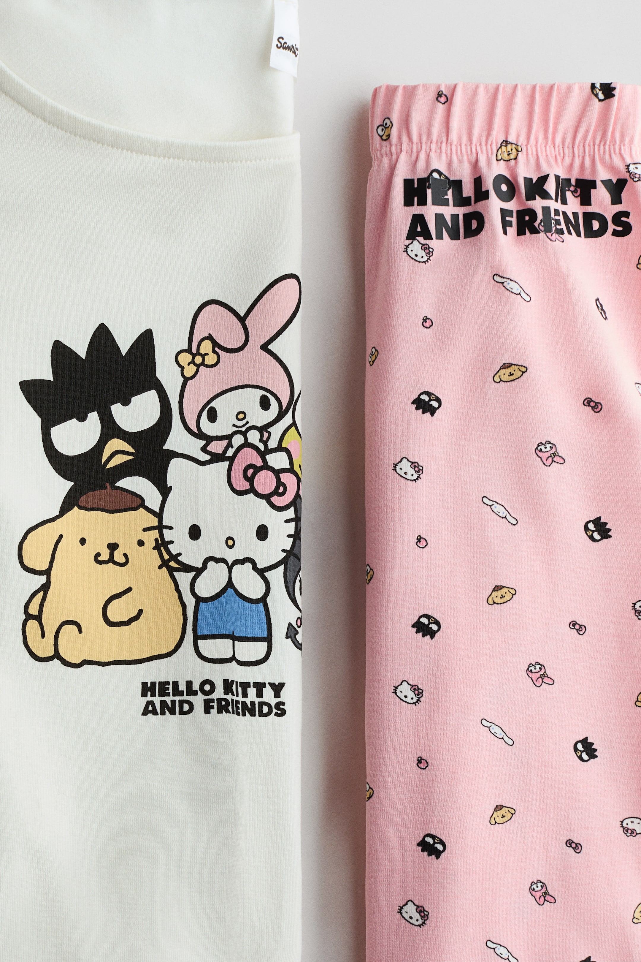 Größeres Bild ansehen: Ein Schlafanzug-Set mit cremefarbenem Oberteil und pinken Hosen. Das cremefarbene Oberteil zeigt einen großen Grafikdruck von Sanrio-Figuren, darunter Badtz-Maru, Pompompurin, My Melody und Hello Kitty auf der Vorderseite, mit dem Text "HELLO KITTY UND FREUNDE" darunter. Die pinken Hosen haben einen elastischen Bund und einen Allover-Print kleinerer Sanrio-Figuren, zusammen mit vertikalem Text "HELLO KITTY UND FREUNDE".
