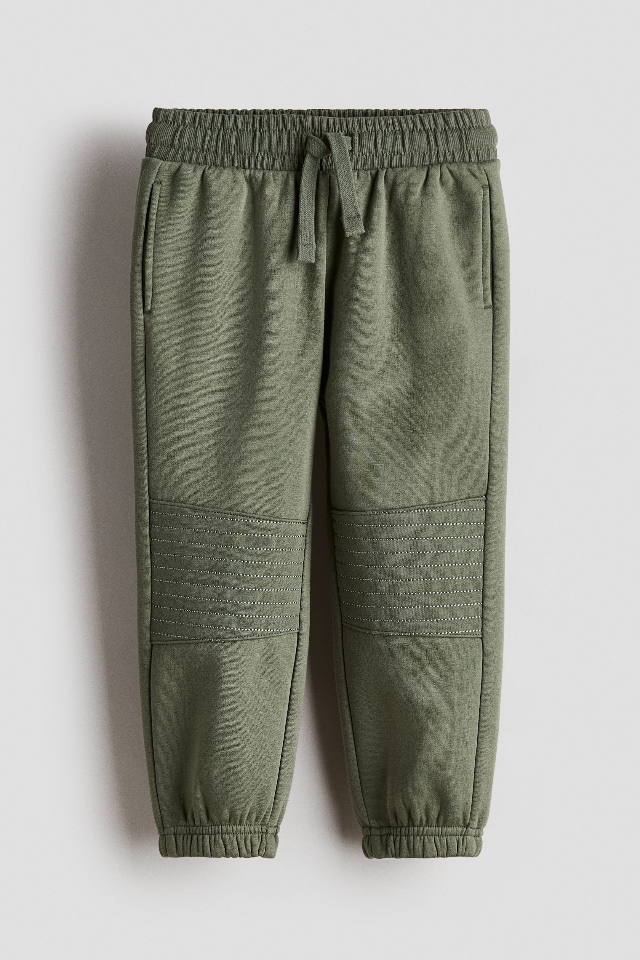 View larger image: A pair of olive green tracksuit bottoms is shown front-on, featuring an elasticated waistband with a drawstring and side pockets. The design includes ribbed knee panels with horizontal stitching and elasticated ankle cuffs.