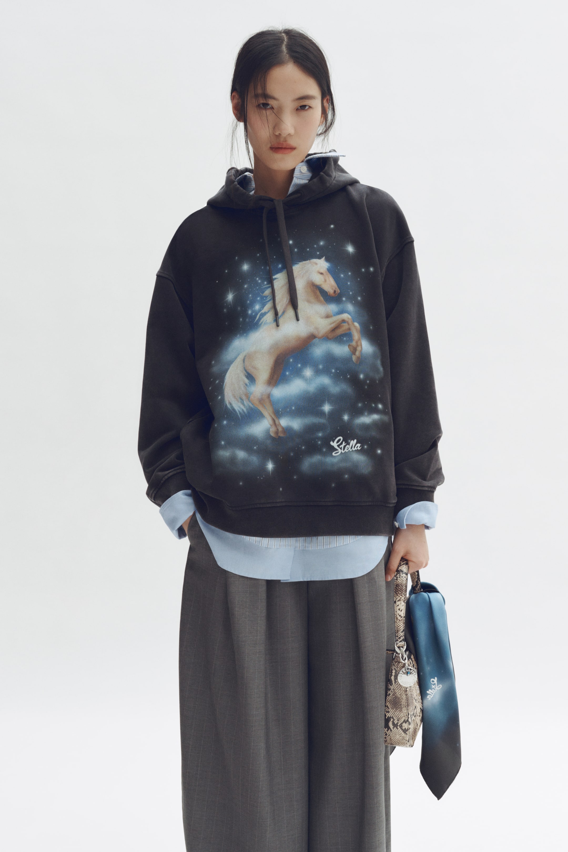 Größeres Bild ansehen: Ein dunkelgrauer Hoodie zeigt ein großes Grafikmotiv eines hellfarbenen Pferdes, das sich vor blauen Wolken und weißen Sternen aufbäumt, mit dem Schriftzug „Stella“ darunter. Dunkle Kordeln ragen aus der Kapuze hervor, und ein hellblaues Hemd mit Kragen ist unter dem Saum und den Bündchen des Hoodies sichtbar.