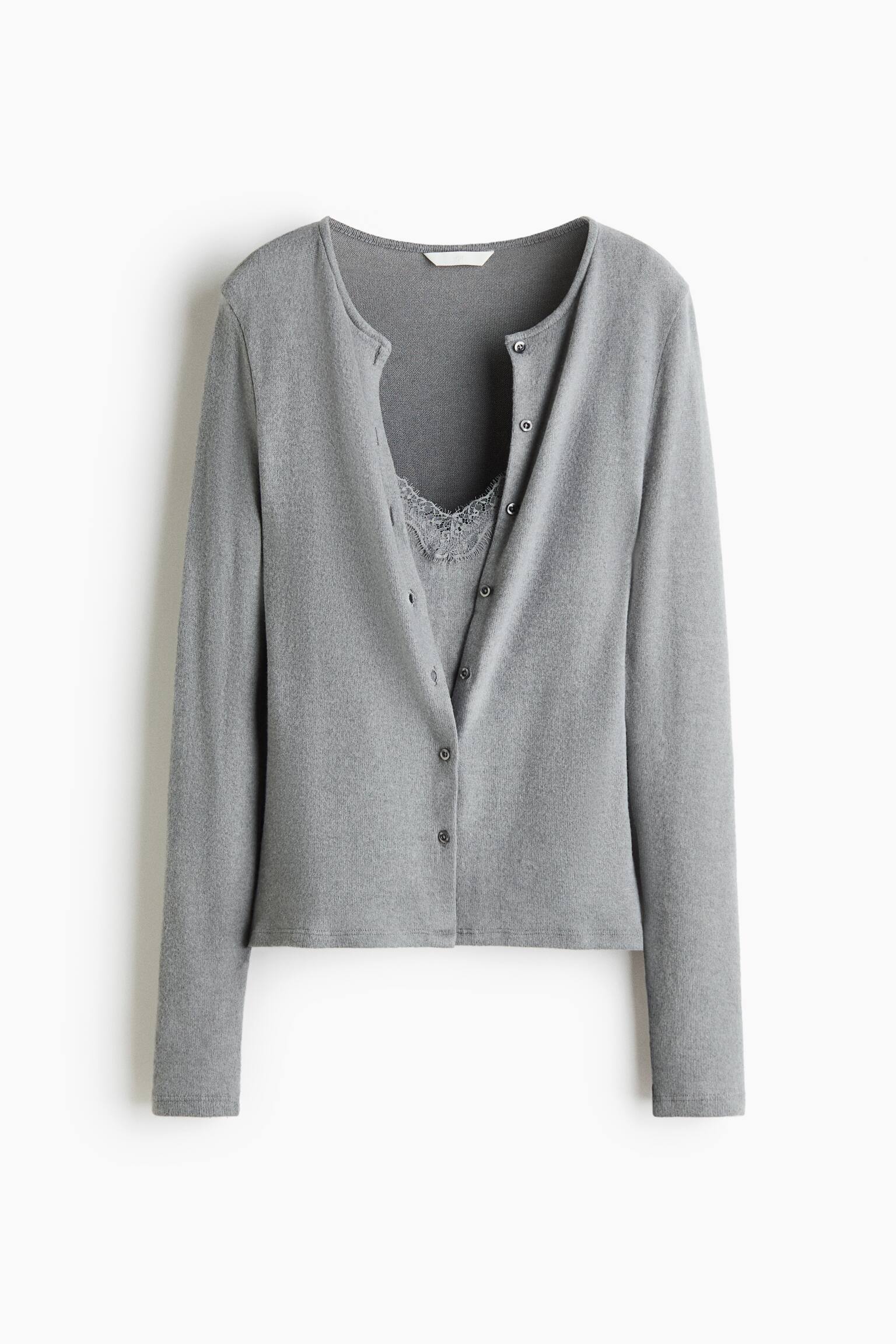 Un gilet en maille gris est prÃ©sentÃ© de face, avec un col rond, des manches longues et une fermeture boutonnÃ©e sur le devant. Le gilet est portÃ© avec les boutons du haut dÃ©faits, laissant apparaÃ®tre un empiÃ¨cement assorti en dentelle bordÃ©e.