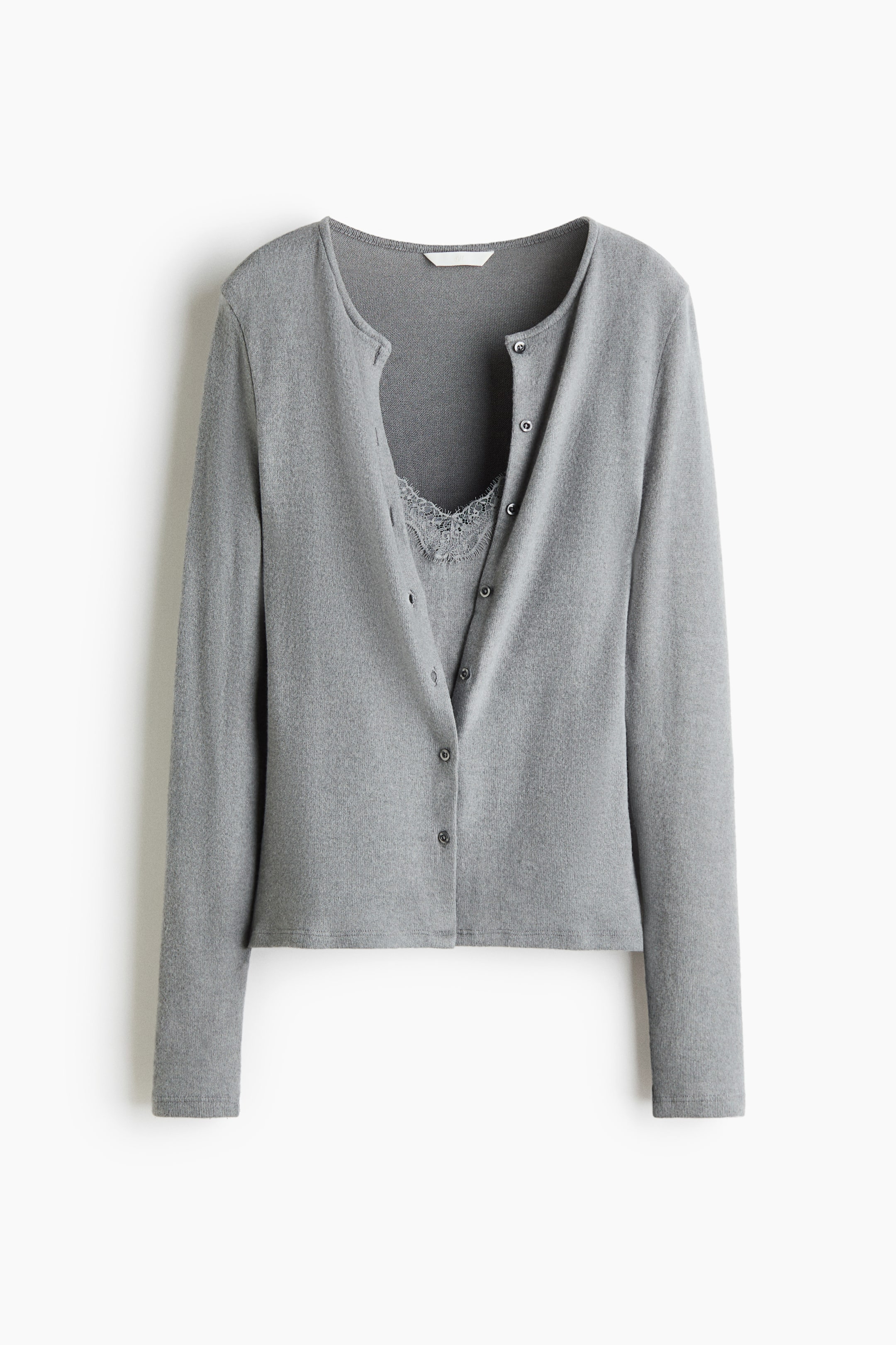 Agrandir l'image: Un gilet en maille gris est présenté de face, avec un col rond, des manches longues et une fermeture boutonnée sur le devant. Le gilet est porté avec les boutons du haut défaits, laissant apparaître un empiècement assorti en dentelle bordée.