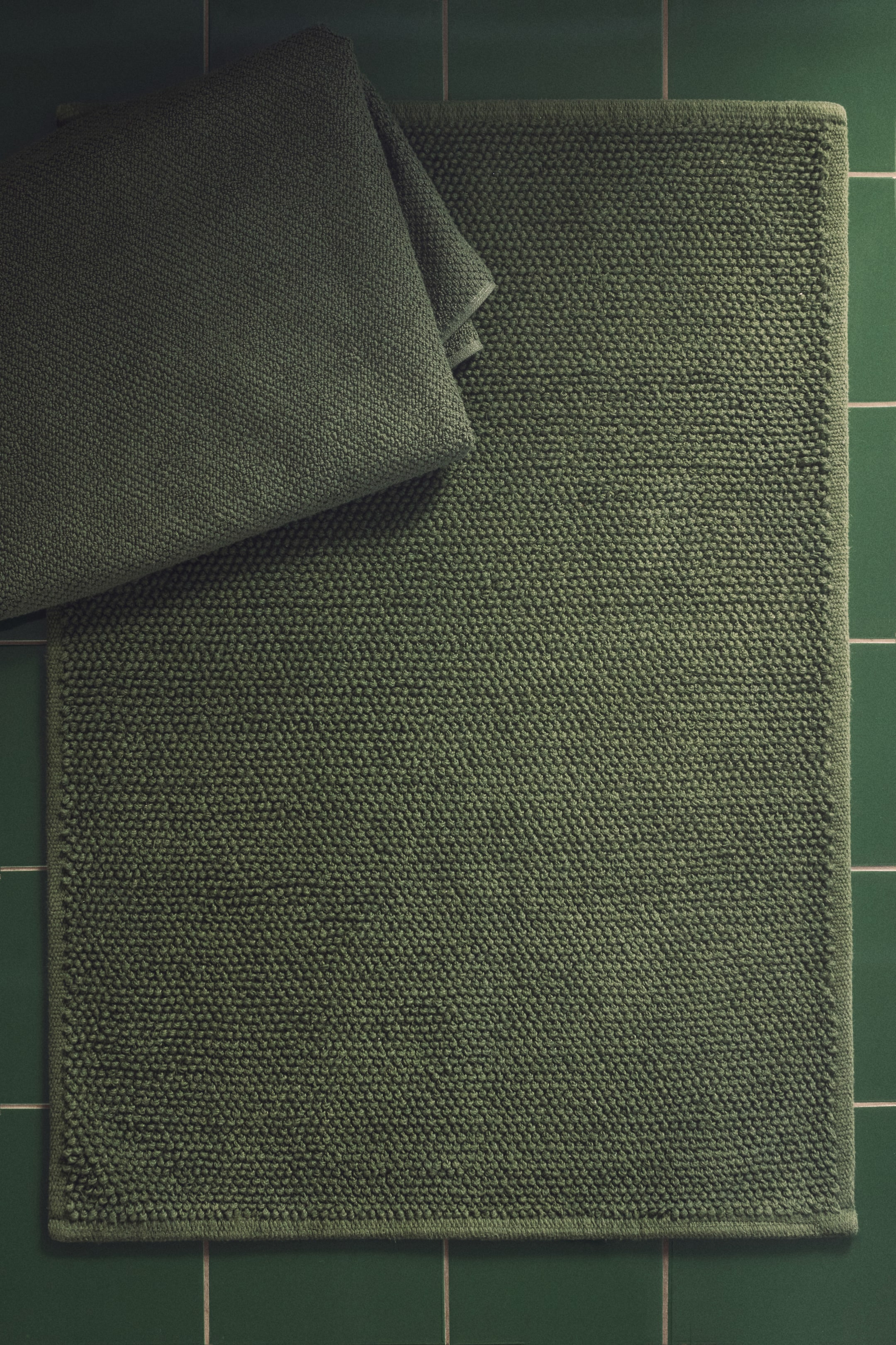 View larger image: A rectangular, deep olive-green bath mat lies flat, displaying a textured bobble surface. A matching deep olive-green towel is folded and rests on the mat's top-left corner.