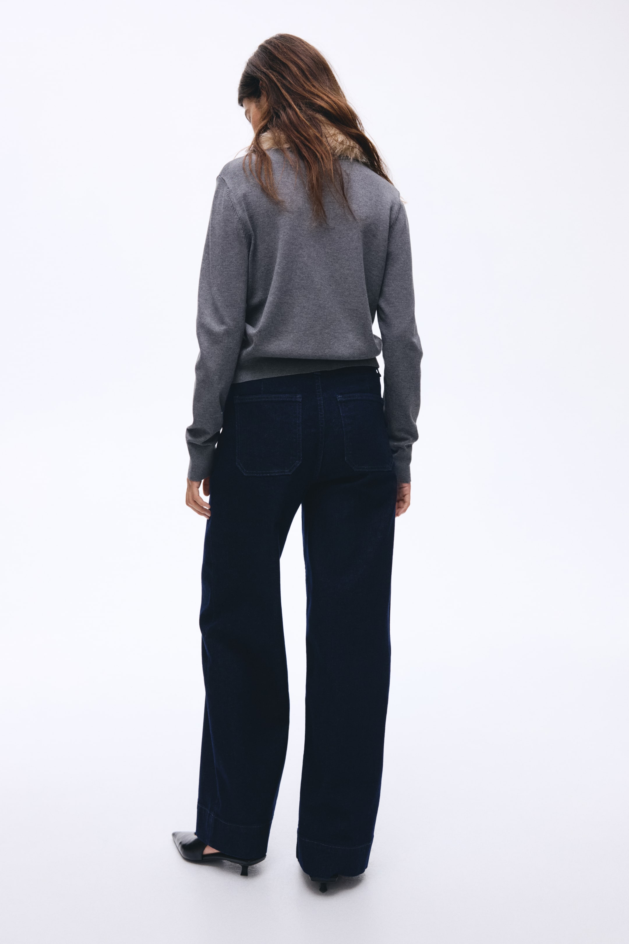 Se større billede: Set bagfra ses en grå langærmet sweater med en lysebrun tekstureret kravekant synlig ved halsudskæringen. Mørkeblå bukser med brede ben har også to store rektangulære patch-lommer.
