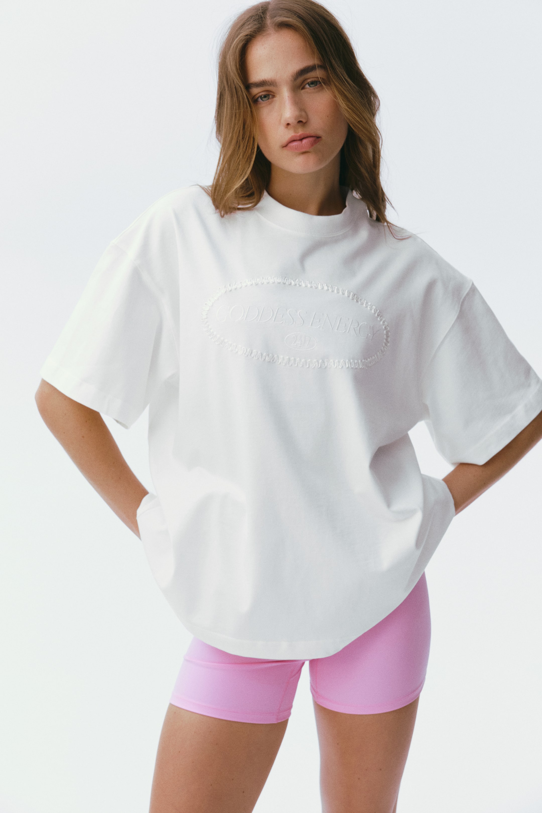 View larger image: A white sports t-shirt is being presented front-facing, featuring a round, ribbed neckline, dropped shoulders, and short sleeves. It showcases an oversized fit with a straight-cut hem and an embossed oval graphic displaying 'GODDESS ENERGY 24/7' on the front chest.