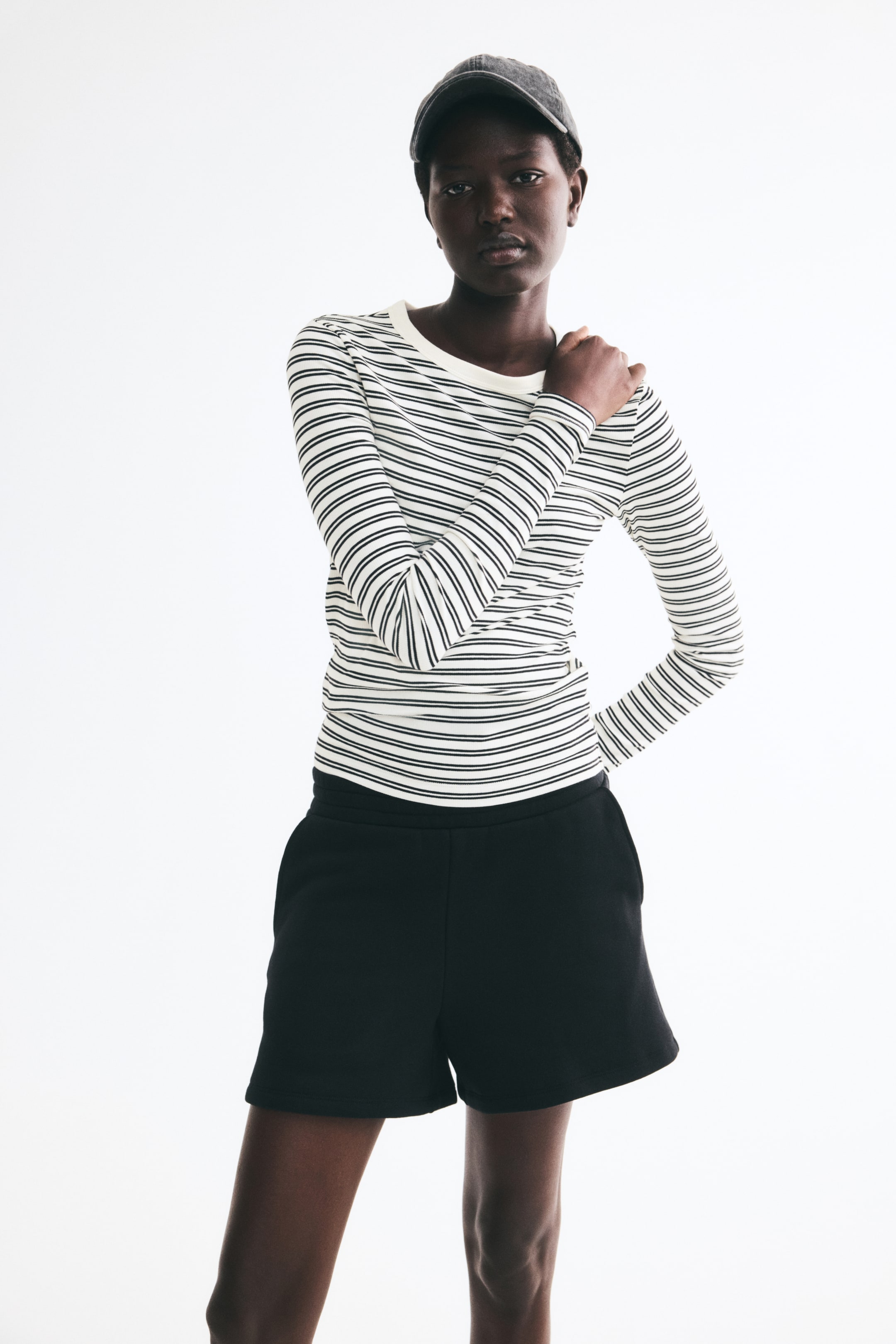 View larger image: A fitted, long-sleeved top featuring black-and-white horizontal stripes, a ribbed texture, and a cream-colored round neckline is being worn by the model. The model is posing with one hand resting on their shoulder, styling the top with black shorts and a dark gray cap.