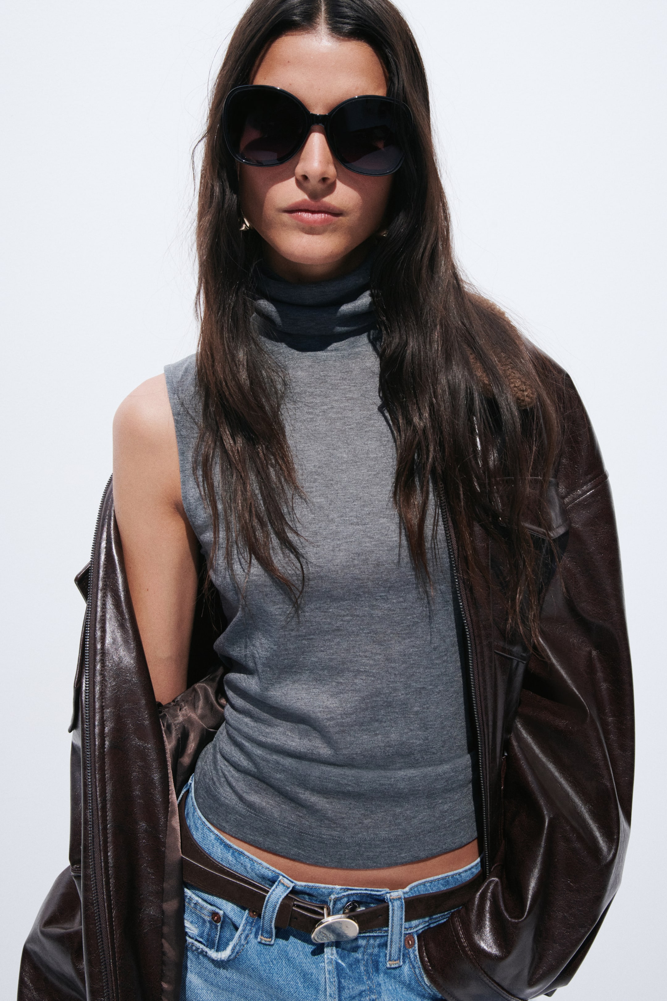 View larger image: A grey sleeveless turtleneck top features a fitted, cropped silhouette. A brown jacket is draped over the shoulders, partially covering the top's sides.