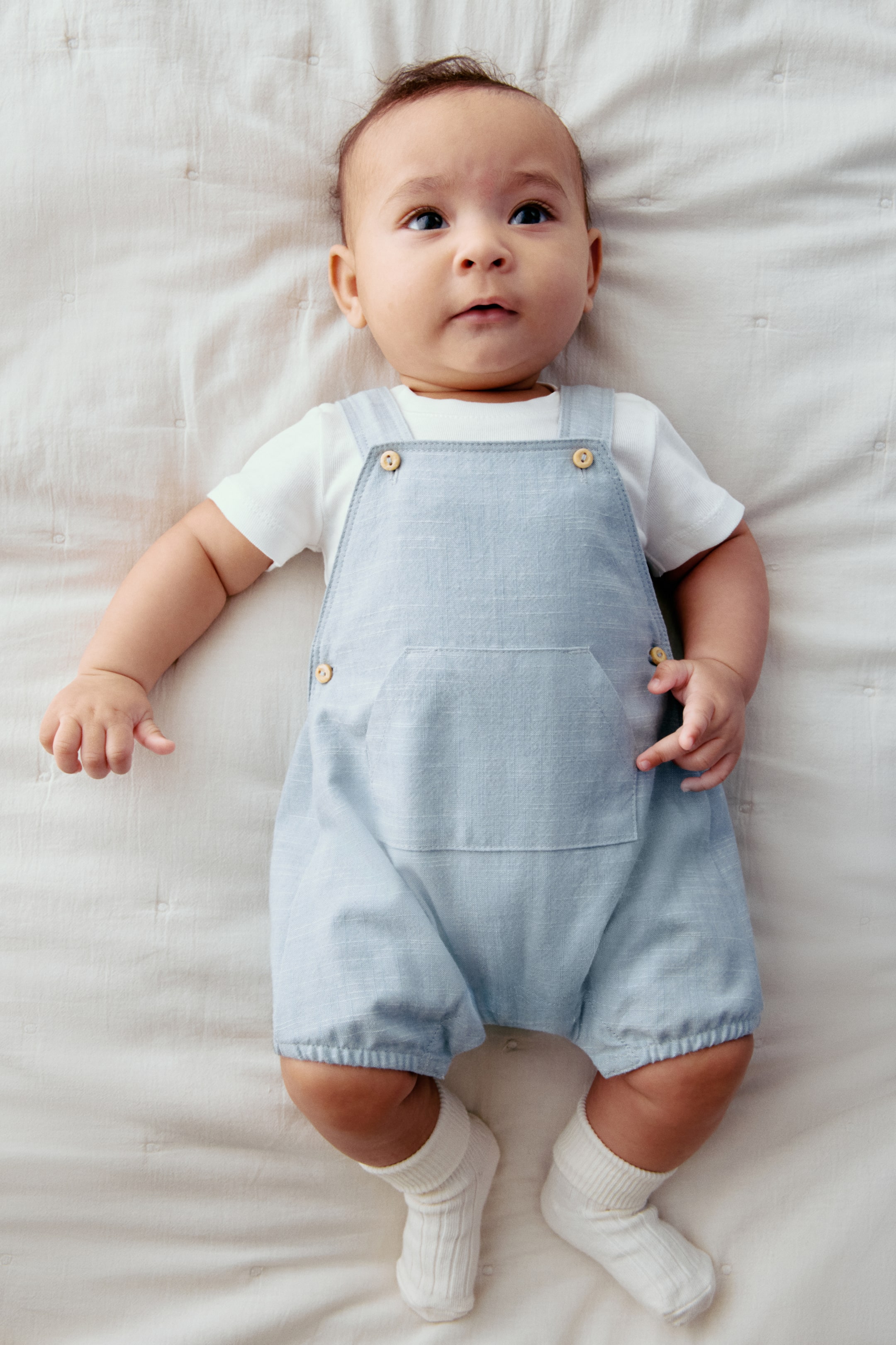 View larger image: A light blue baby romper is being worn, featuring a large patch pocket on the front and wooden buttons on the shoulder straps. It is being layered over a white short-sleeved t-shirt, and white ribbed socks are completing the outfit.