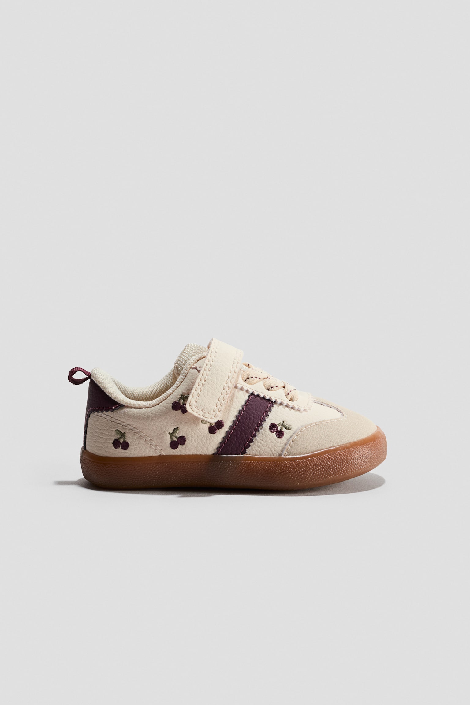 A barefoot trainer is resting in a side-profile view, featuring an off-white textured leather upper adorned with embroidered cherry details and a dark purple diagonal stripe. The shoe is showcasing a hook-and-loop strap closure over elastic laces and a flexible gum rubber sole that wraps around the toe and heel.