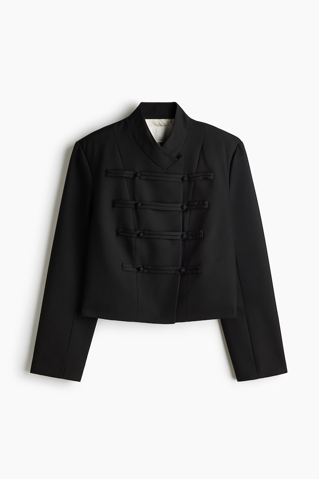 A black cropped jacket is facing front, featuring a structured, high wrap-over collar and long, straight sleeves. The front is displaying four horizontal rows of fabric-covered toggle loop closures. - 1