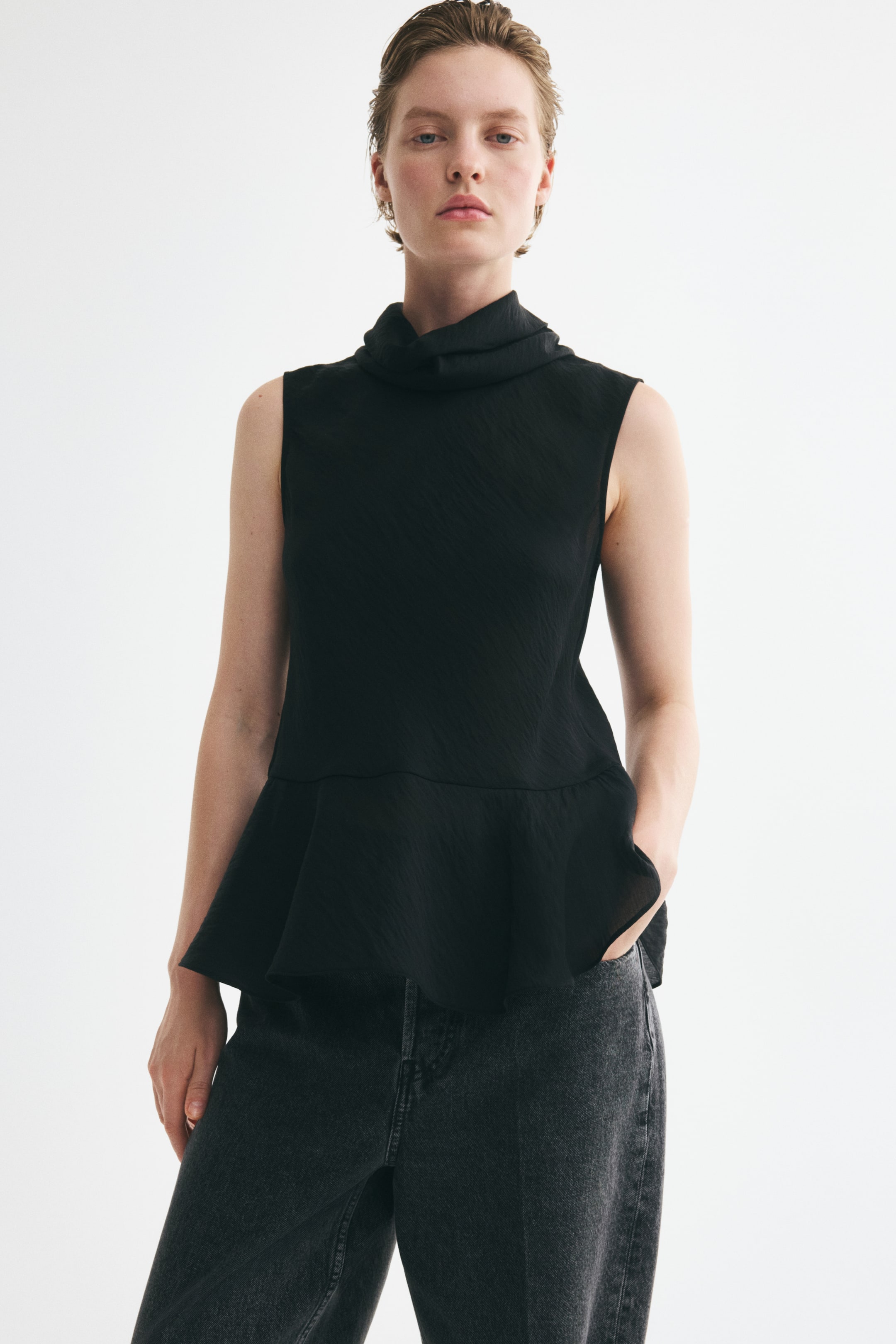View larger image: A black long sleeveless top, featuring an airy weave, a roll neck, and a flared peplum, is being worn by the model. The model is standing, with one hand in the pocket of dark trousers worn with the top.
