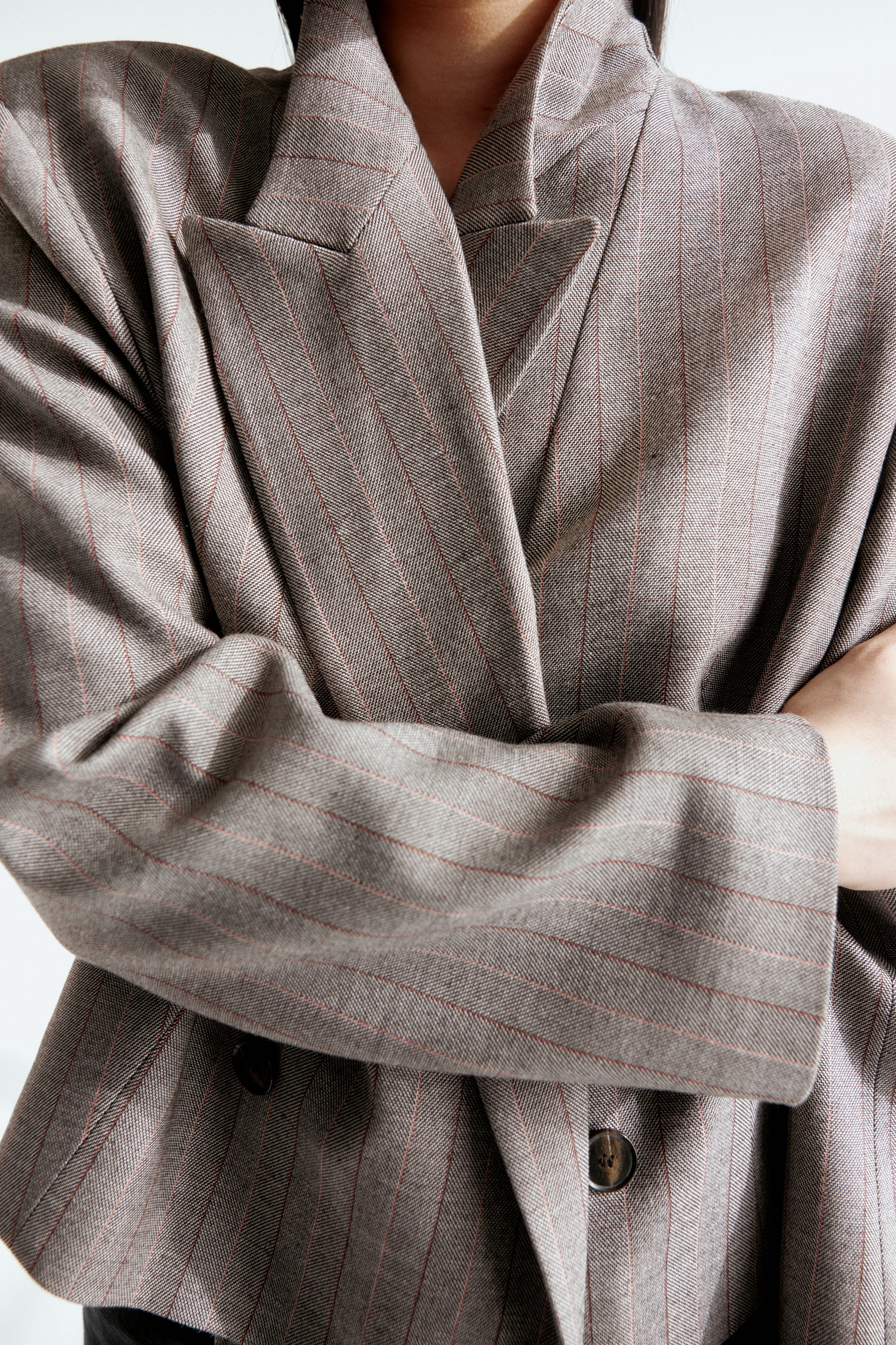 Grotere afbeelding bekijken: De grijs-bruine krijtstreep blazer heeft brede inkeping revers, met een subtiele visgraatweving en dunne rode krijtstrepen overal. Een donkerbruine knoop is zichtbaar op de voorste sluiting, en de ontspannen pasvorm van de mouwen valt met natuurlijke plooien door de gekruiste armen van de drager.