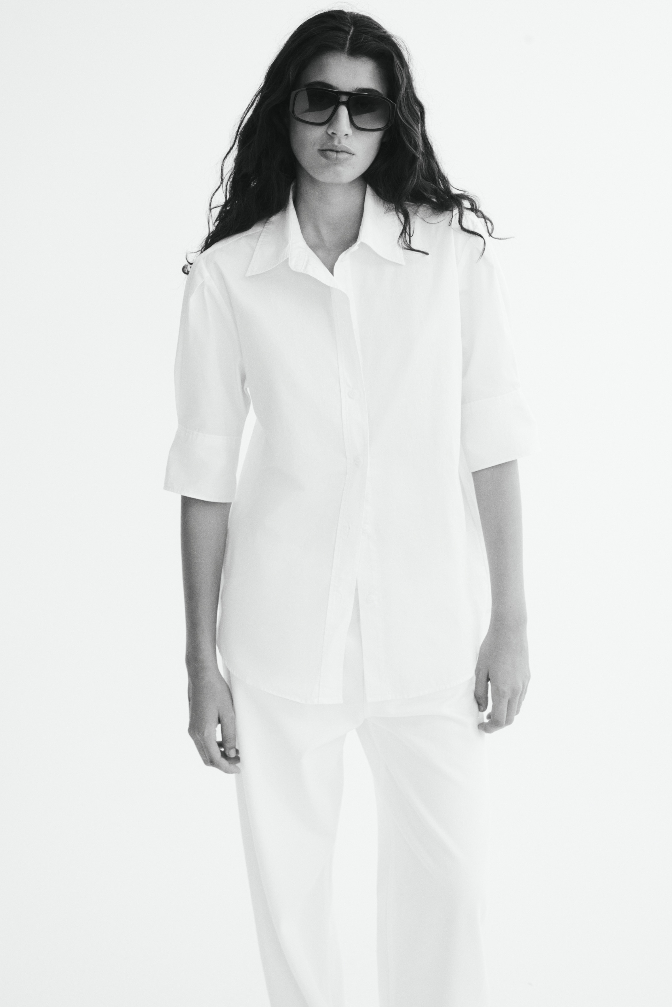 A white cotton poplin shirt is shown from the front, featuring a classic pointed collar, a button-down front, and wide, elbow-length sleeves with subtle pleat details. The shirt is hanging flat, showing a relaxed silhouette and a rounded hem.