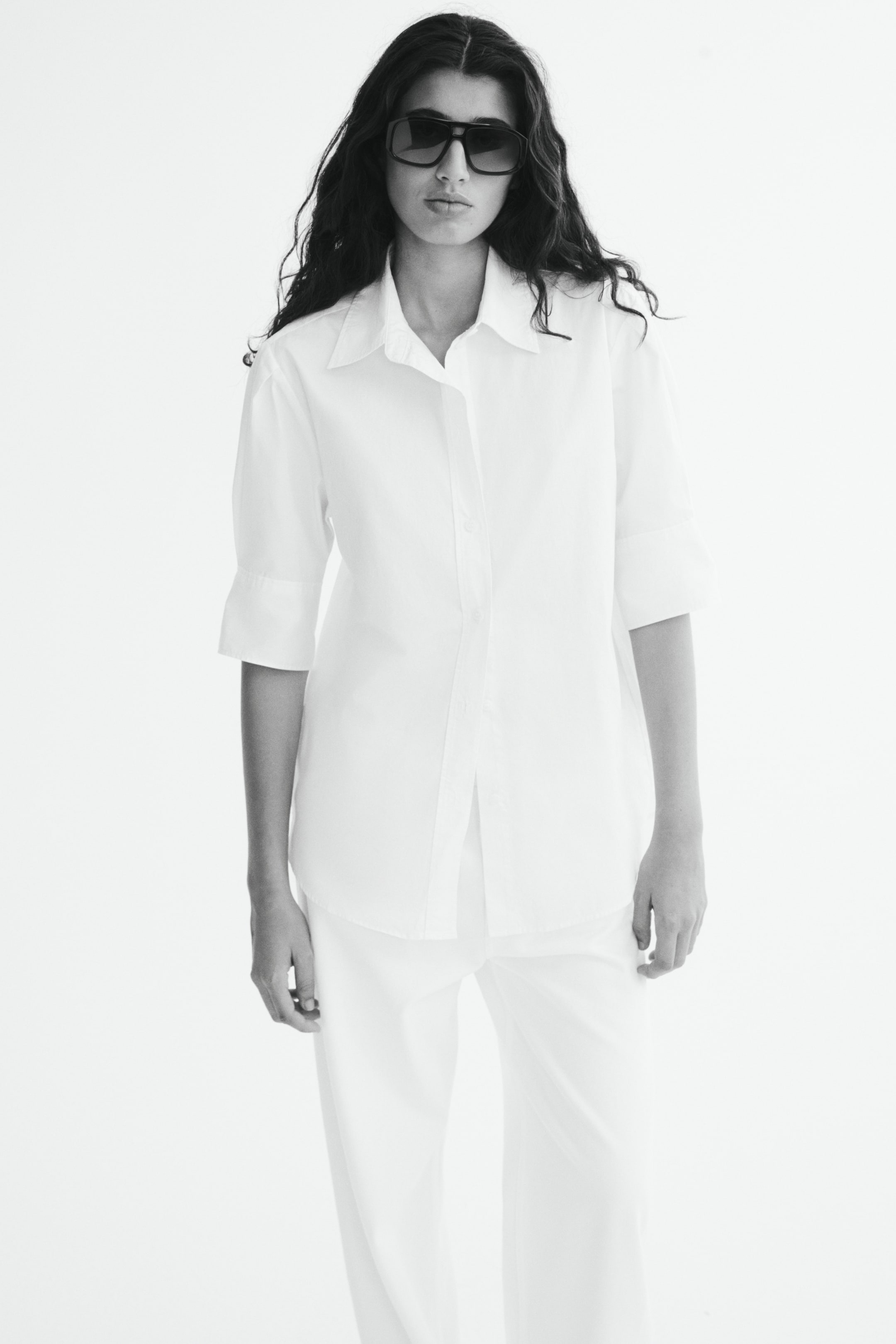 View larger image: The model is wearing a white cotton poplin shirt featuring a classic collar, a button-down front, and elbow-length sleeves with wide cuffs. The shirt is styled loosely over matching white trousers.