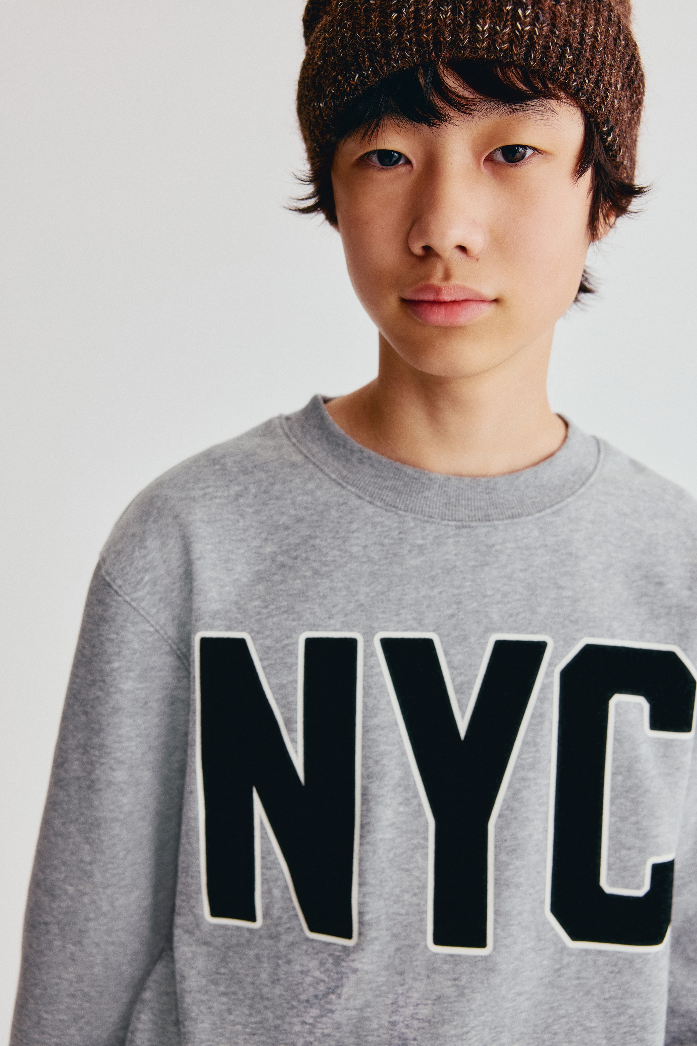 Una sudadera gris muestra su parte frontal, con "NYC" en grandes letras negras con un contorno blanco a lo largo del pecho. Está diseñada con un cuello redondo acanalado, puños acanalados y un dobladillo acanalado.