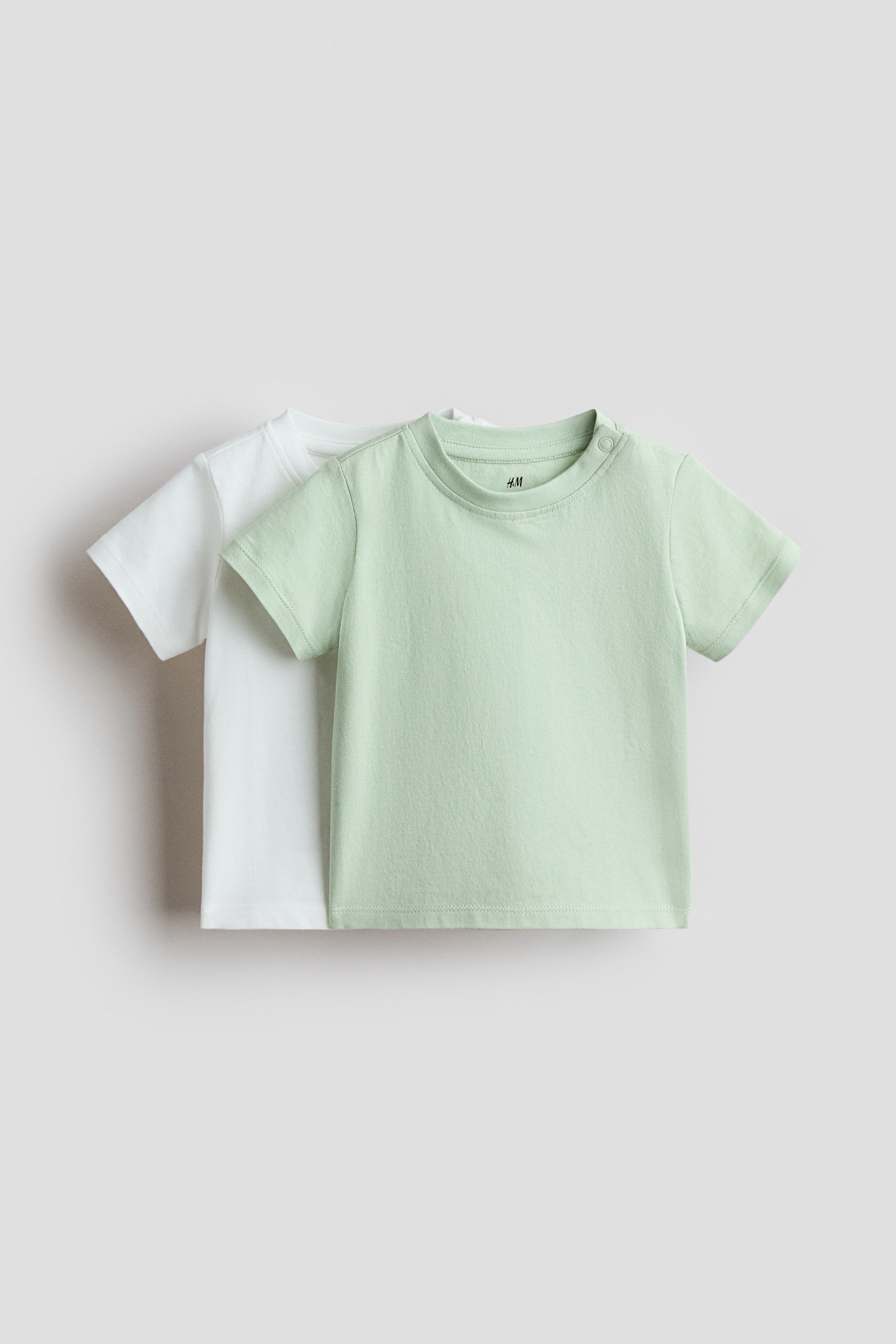 View larger image: Introducing a 2-pack of cotton T-shirts, showcasing one light green and one white short-sleeved top. The light green T-shirt, displayed front-facing, features a round neck and two snap buttons on the left shoulder, partially concealing the white T-shirt layered beneath.