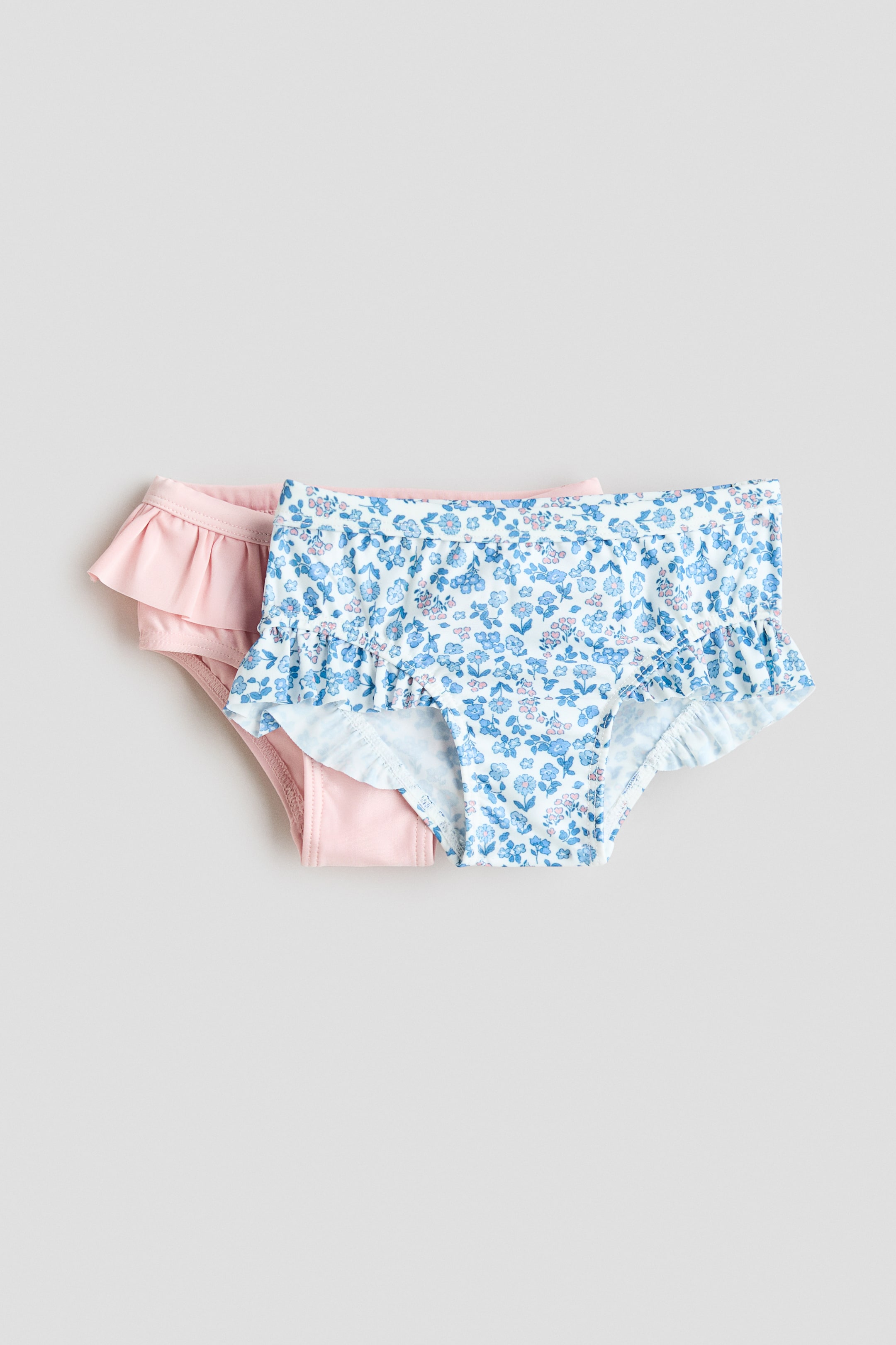 Agrandir l'image: Deux culottes de bain à volants sont présentées de face, l'une en rose clair uni et l'autre avec un fond blanc orné d'un motif floral bleu et rose. Les deux culottes sont dotées d'une élastication étroite à la taille et d'un gousset doublé.