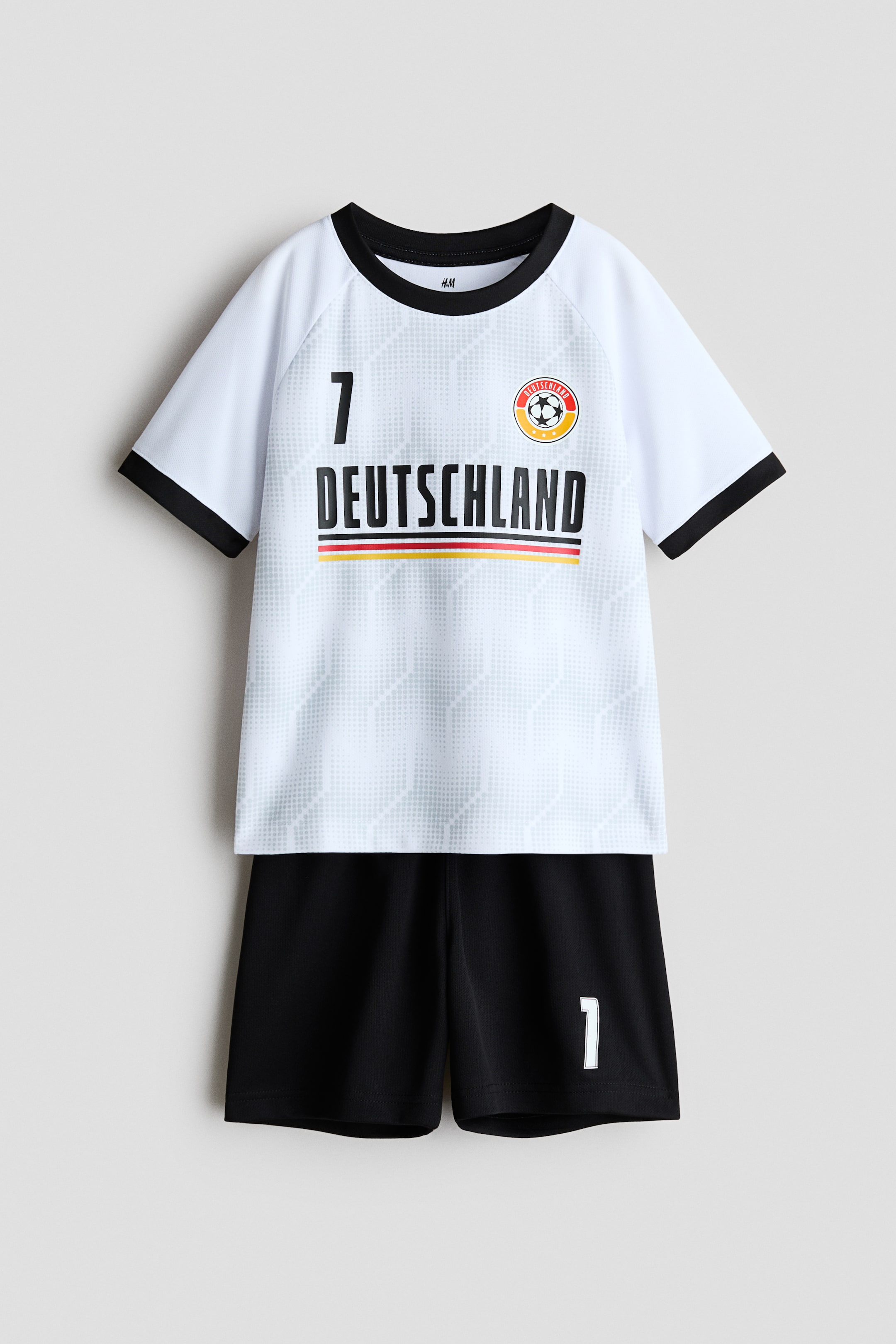 Agrandir l'image: Un ensemble de football imprimé est présenté de face, avec un haut à manches courtes blanc en tissu mesh doux, doté d'un col et de poignets noirs. Le haut est imprimé avec un motif géométrique subtil, le numéro '7', le mot 'DEUTSCHLAND' au-dessus d'une bande noire, rouge et or, ainsi qu'un écusson de football ; le short noir assorti est doté d'une taille élastiquée avec cordon de serrage et d'un numéro '1' blanc sur le bas de la jambe gauche.