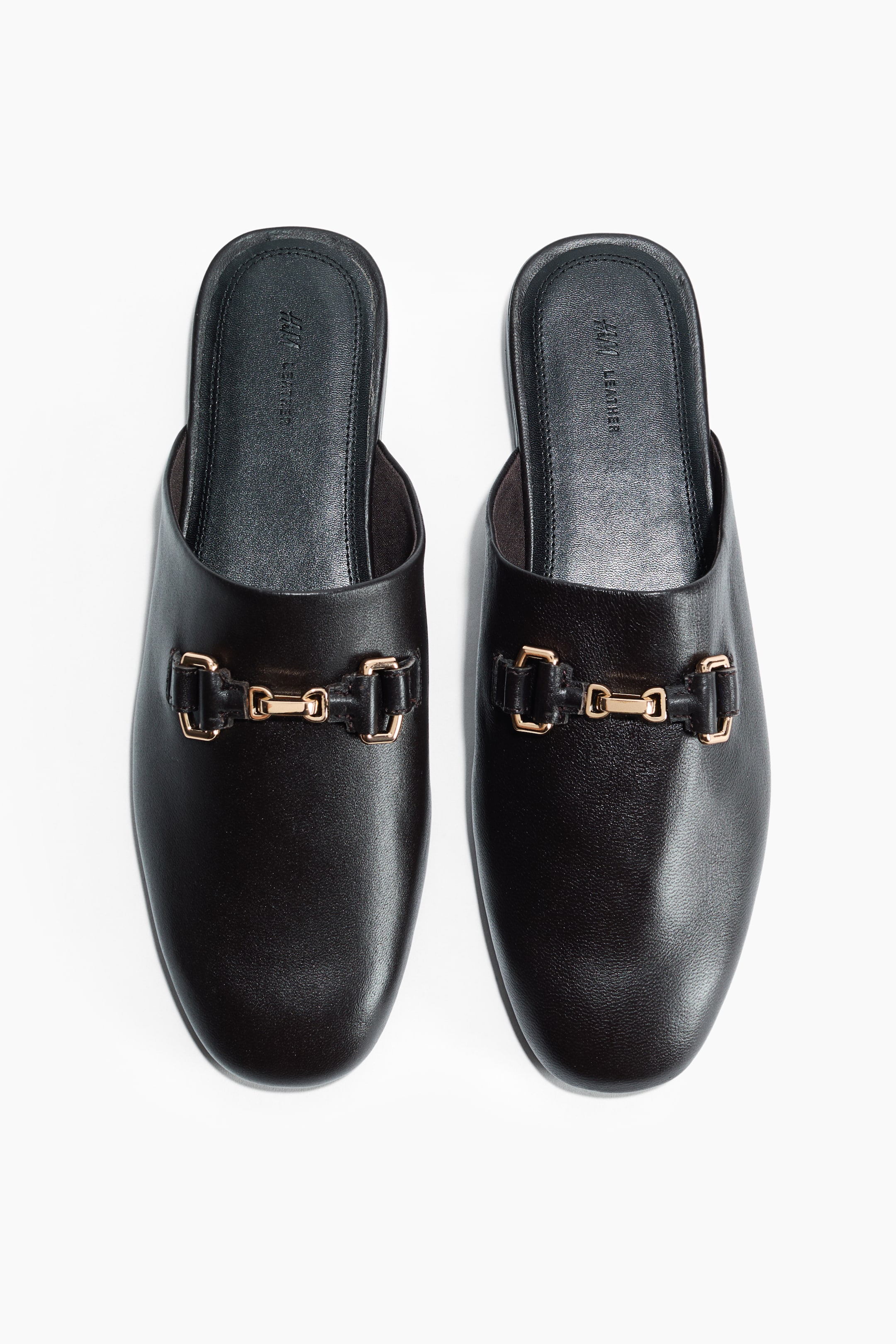 Grösseres Bild ansehen: Ein Paar schwarze Ledermules zeigt ein goldfarbenes Horsebit-Schnallendetail über dem Rist jedes Schuhs. Die schwarzen Innensohlen tragen das Branding "H&M LEATHER".