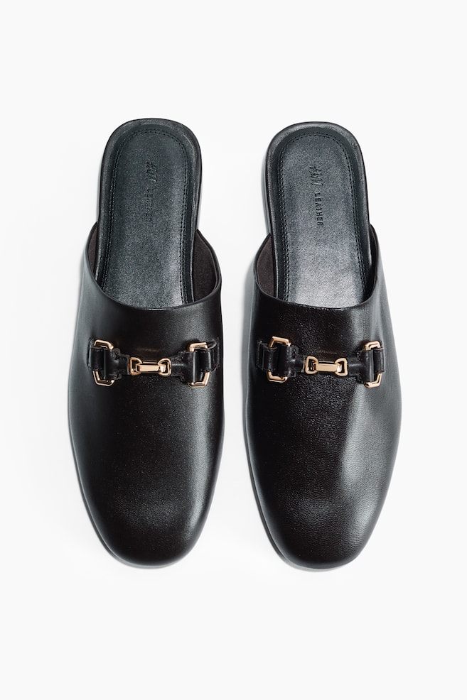 A pair of black leather mules is displaying a gold-toned horsebit buckle detail across the vamp of each shoe. The black insoles are featuring "H&M LEATHER" branding. - 1