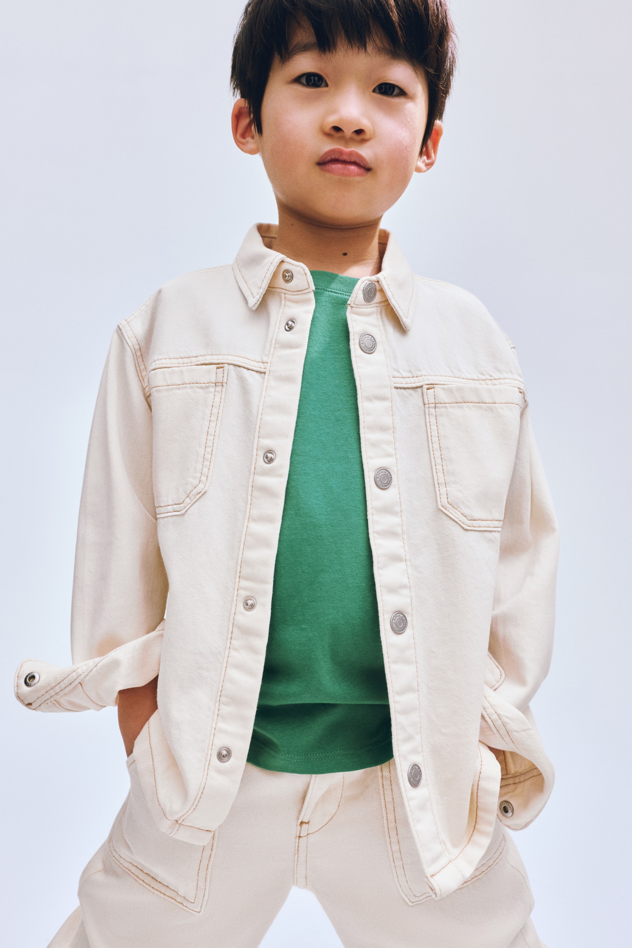 Προβολή μεγαλύτερης εικόνας: Μια τζιν ζακέτα σε κρεμ απόχρωση, με αντίθετες καφέ ραφές, ασημί κουμπιά τύπου σούστα, μυτερό γιακά, δύο τσέπες με καπάκι στο στήθος και δύο κάτω τσέπες με πατ, φοριέται ανοιχτή πάνω από ένα μονόχρωμο πράσινο μπλουζάκι με στρογγυλή λαιμόκοψη. Η μία μανσέτα του μανικιού είναι γυρισμένη και ταιριαστά κρεμ τζιν παντελόνια με αντίθετες καφέ ραφές είναι ορατά από κάτω.