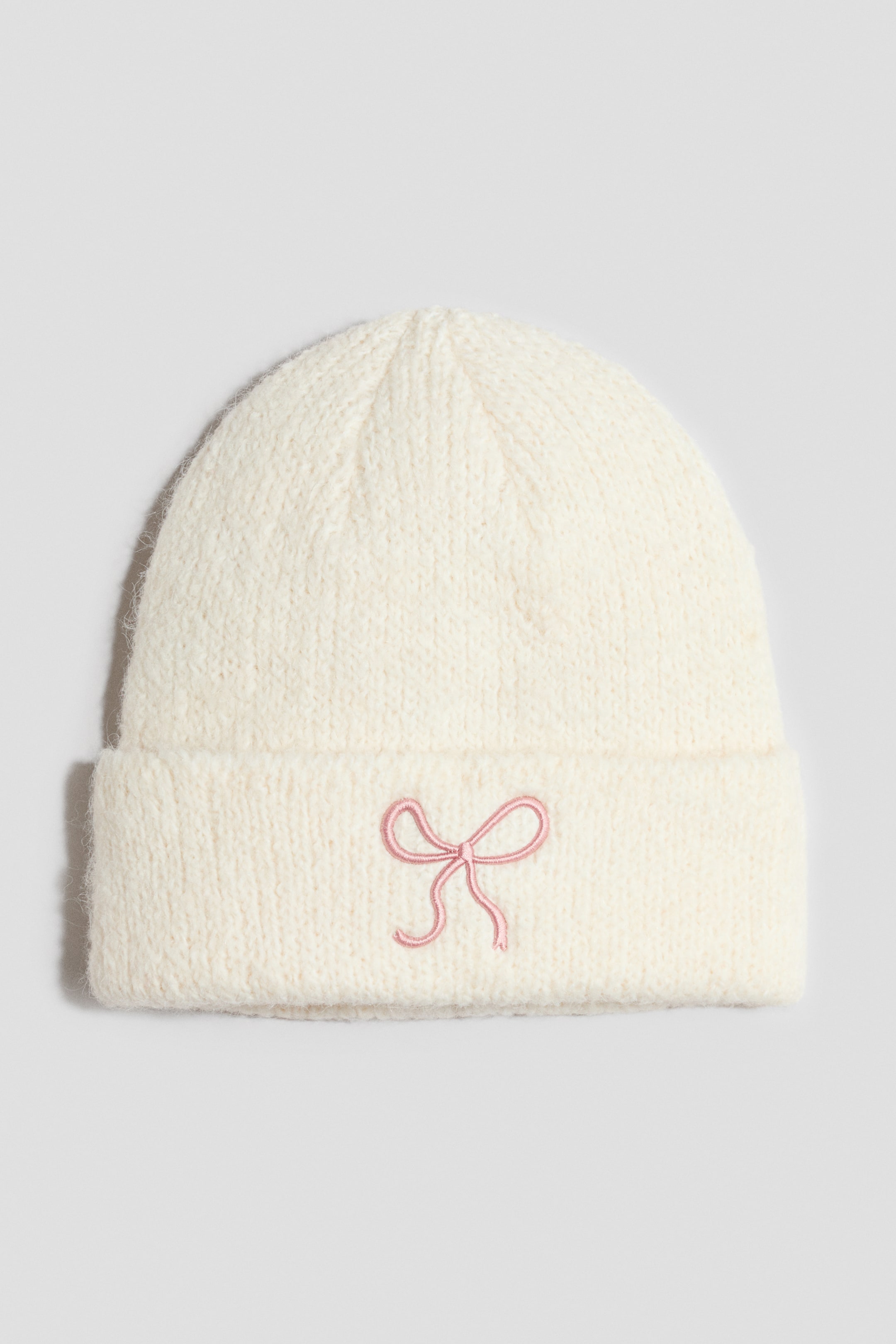 Grösseres Bild ansehen: Eine cremefarbene Beanie aus weichem, flauschigem Strick ist von vorn zu sehen und verfügt über einen breiten Umschlagrand. In der Mitte des umgeschlagenen Saums ist eine kleine, rosa Stickschleife als Detail angebracht.
