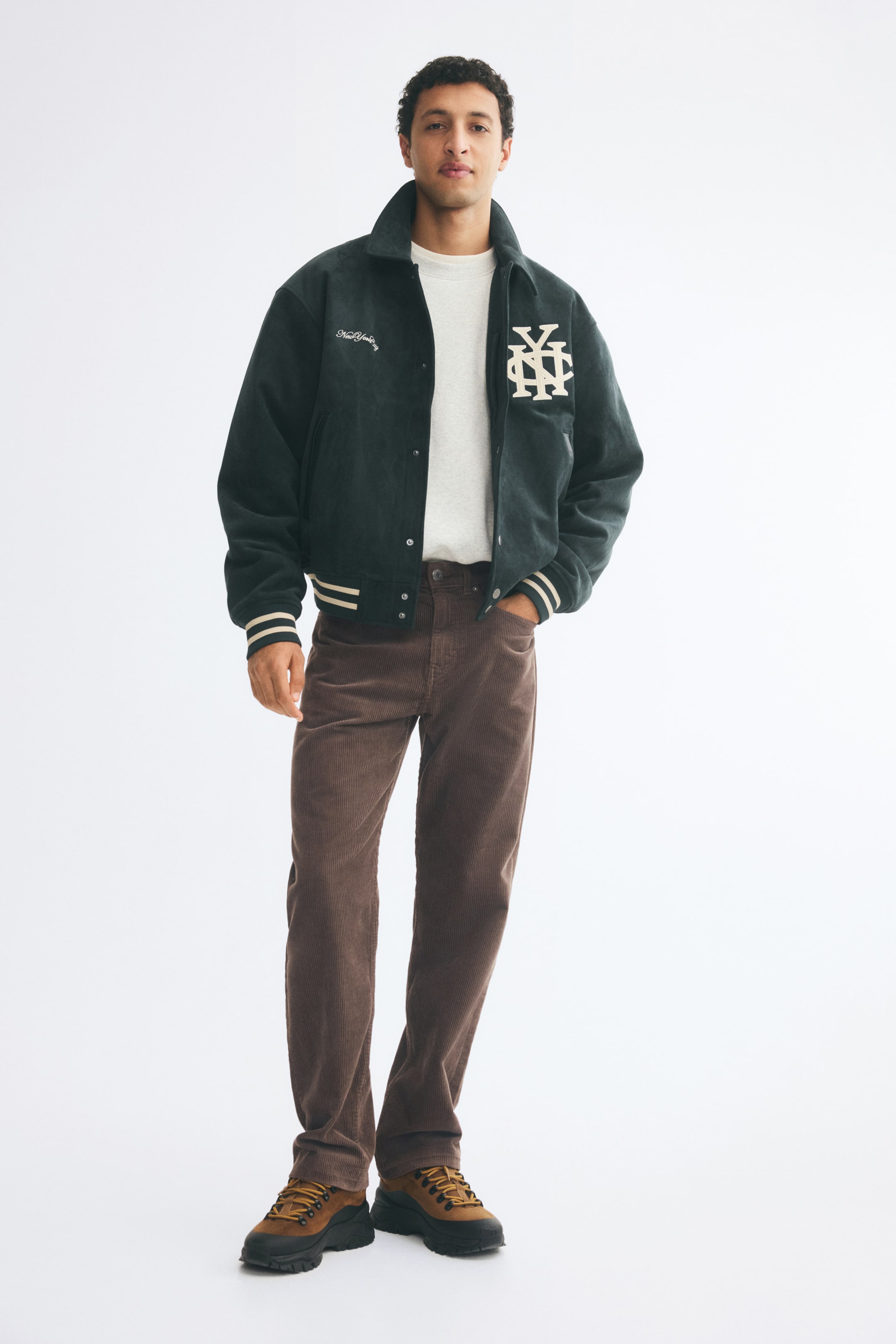 View larger image: Brown, regular-fit cotton corduroy trousers with straight legs are being worn by the model, who is standing with one hand resting in the front pocket. The trousers are styled with a white T-shirt, a dark green varsity jacket, and brown lace-up shoes.